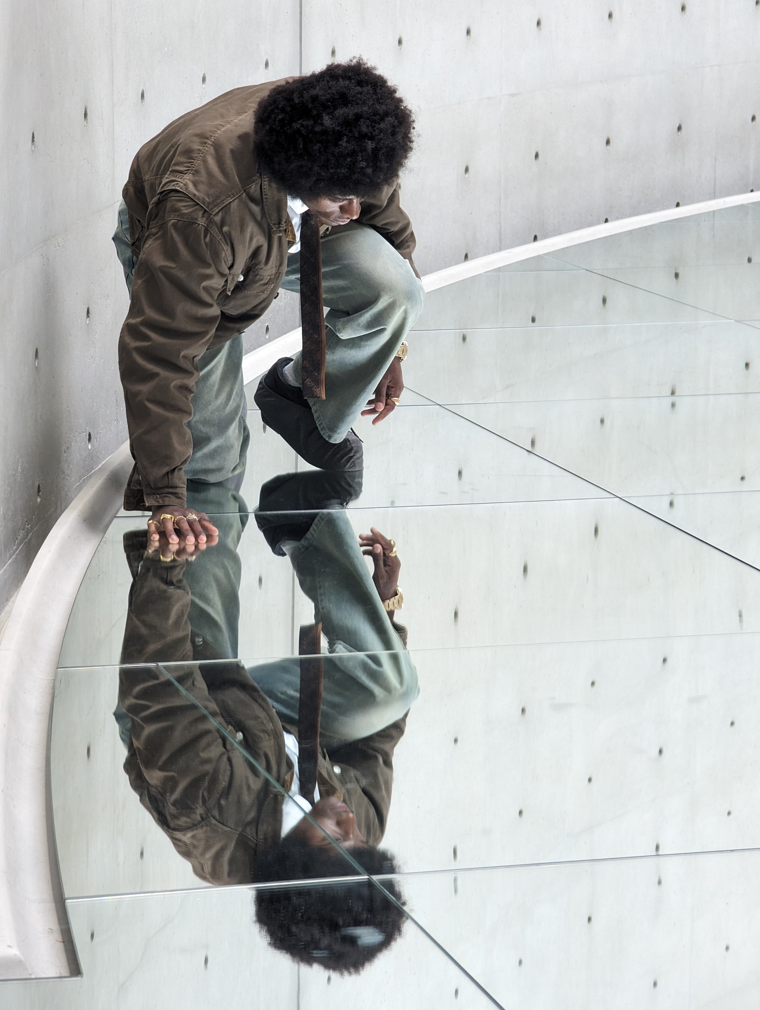 Person with afro hairstyle crouching in front of a concrete wall, reflecting in a mirror, with visible nails on the wall.