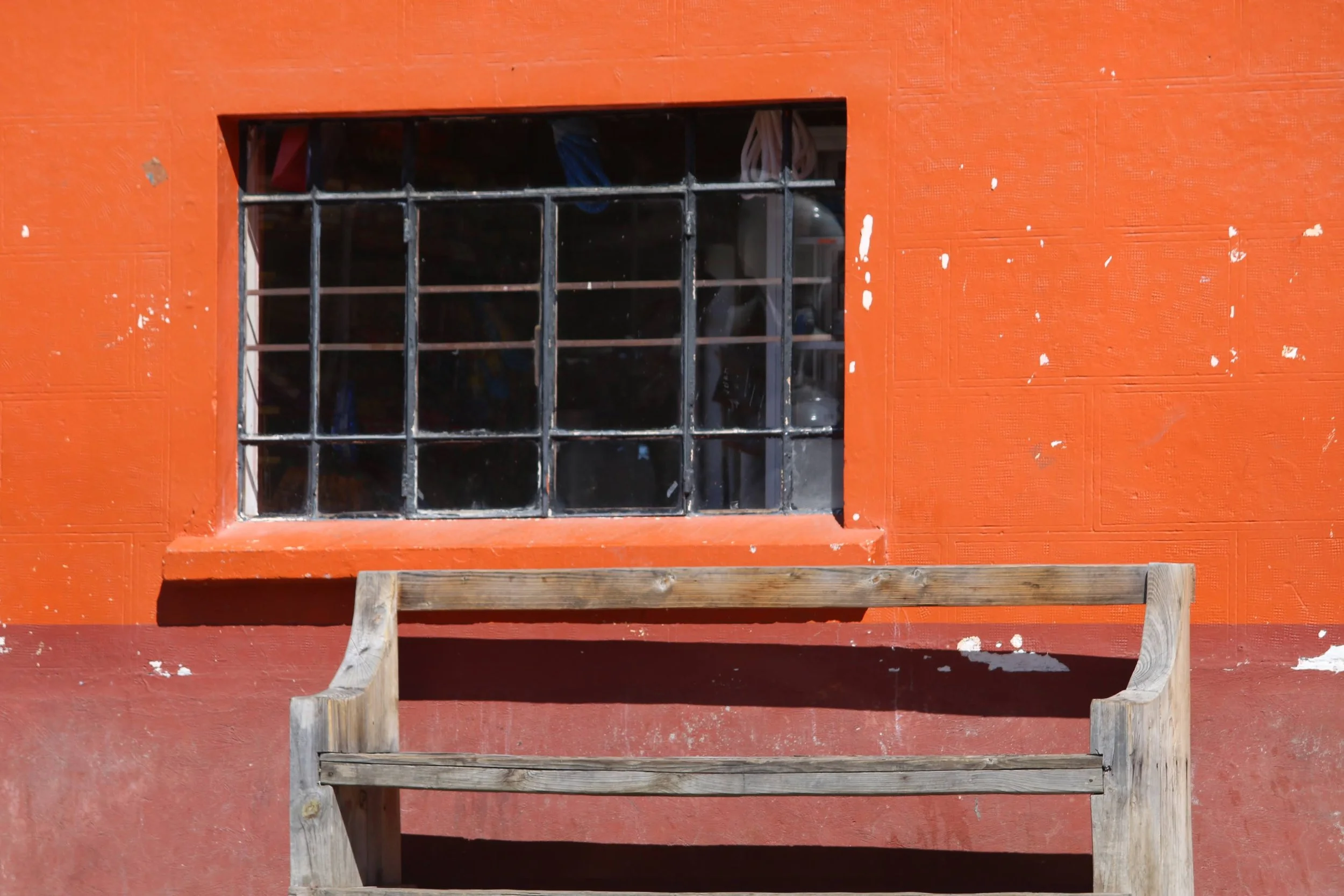 A small window with black metal bars on a red wall, with some wood steps beneath and a wooden platform in front.