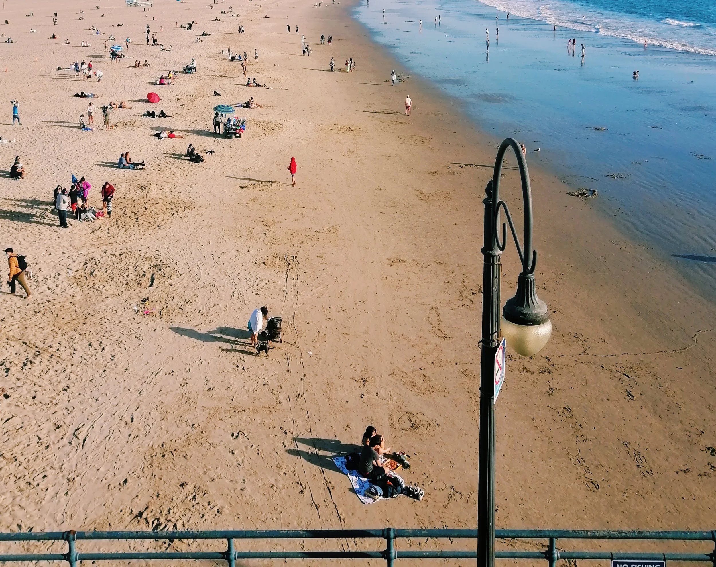A crowded beach with people lounging on towels, walking by the shoreline, and swimming in the water. There are umbrellas and a lifeguard stand visible, along with a metal guardrail in the foreground.