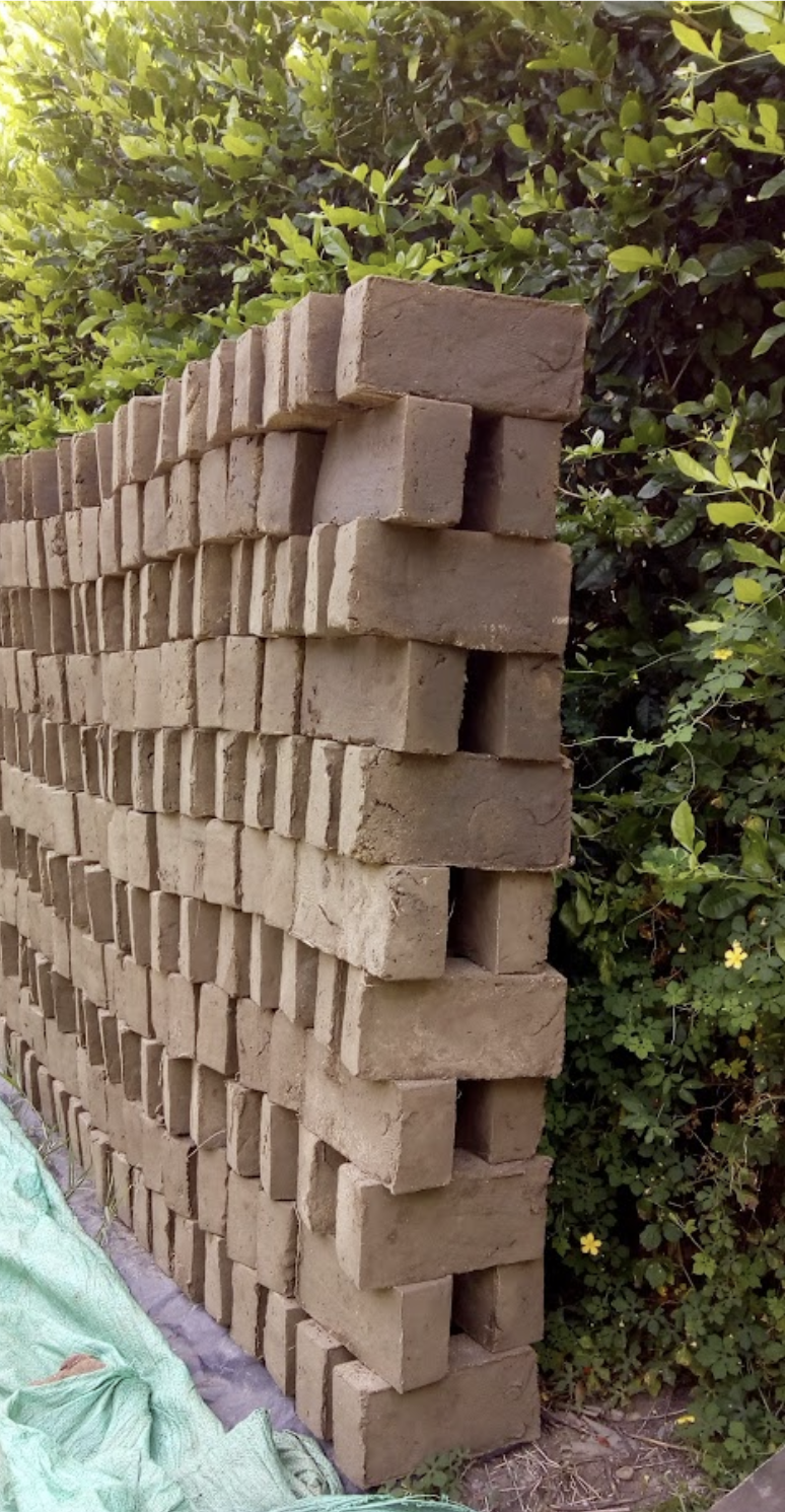 A stack of beige bricks arranged in a staggered pattern outdoors near green bush leaves.