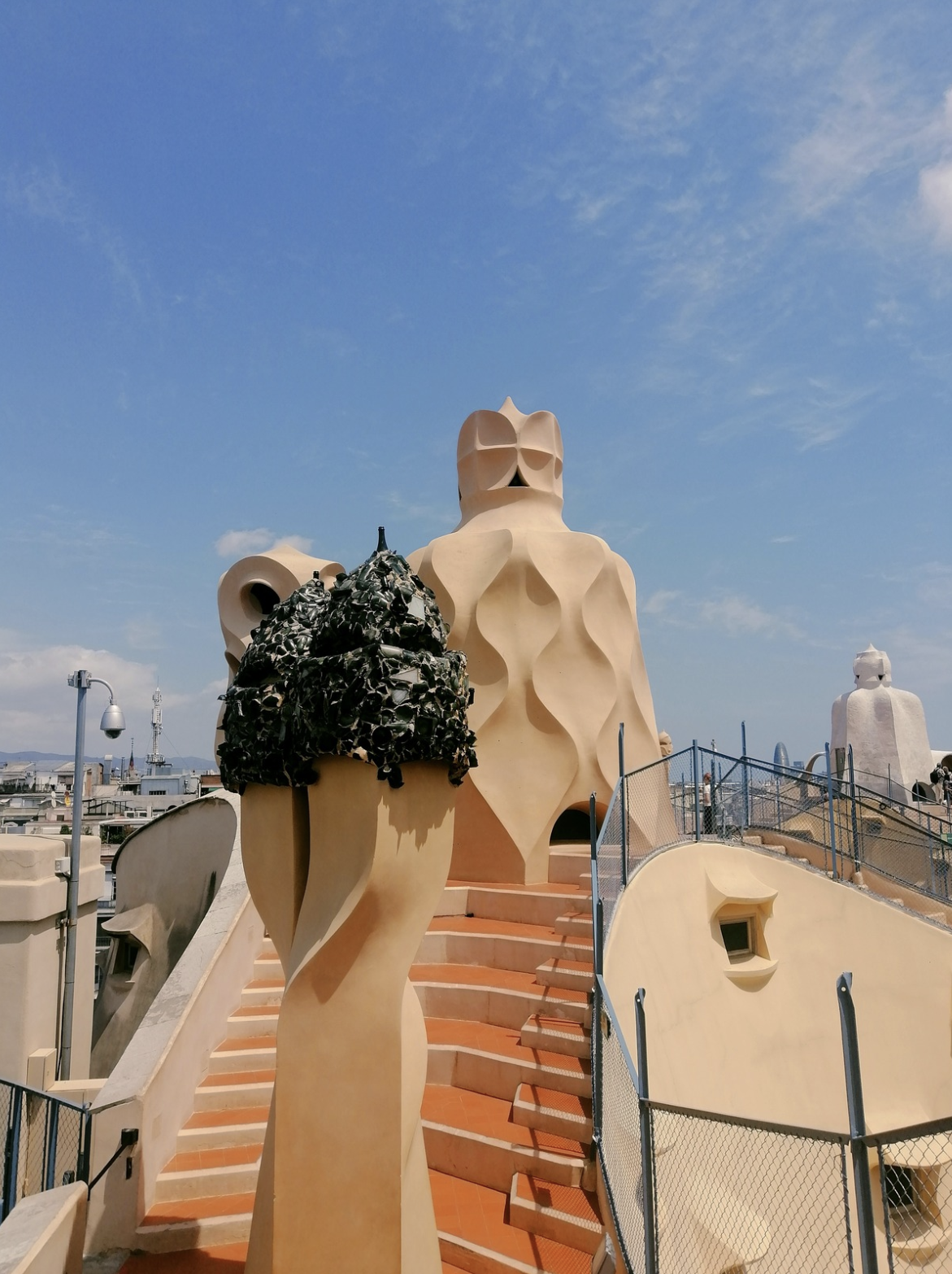 Architectural view from the rooftop of Casa Batlló in Barcelona, Spain, showcasing its distinctive organic shapes, colorful tile work, and sculptures, under a blue sky.