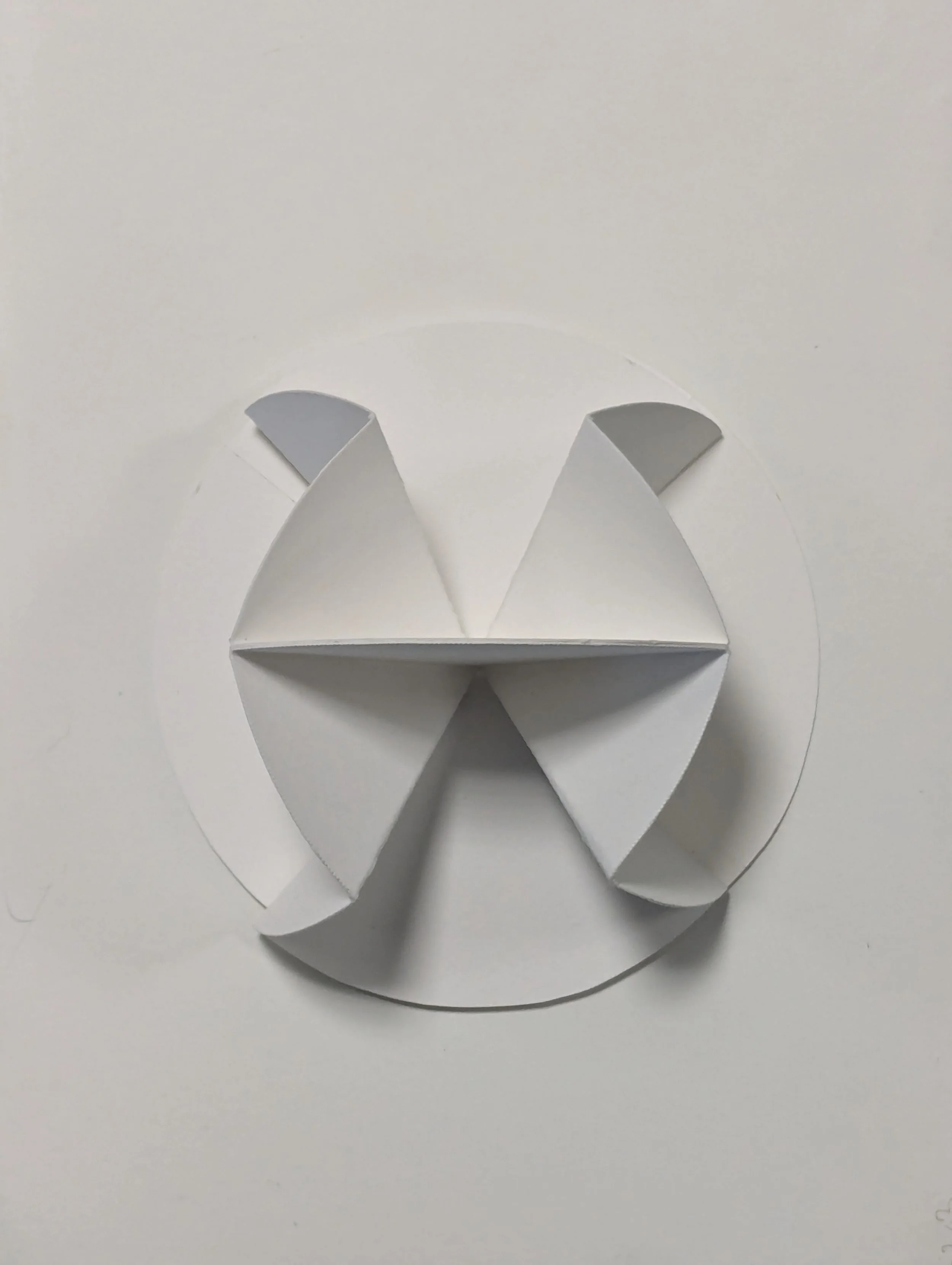 A white paper cutout paper sculpture resembling a cat's face, with pointed ears and a rounded face, placed on a white circular paper background.