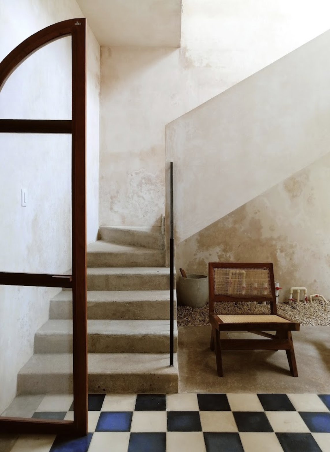 Interior of a minimalist home with a staircase, a wooden chair, a concrete pot, and a checkered black and white floor.