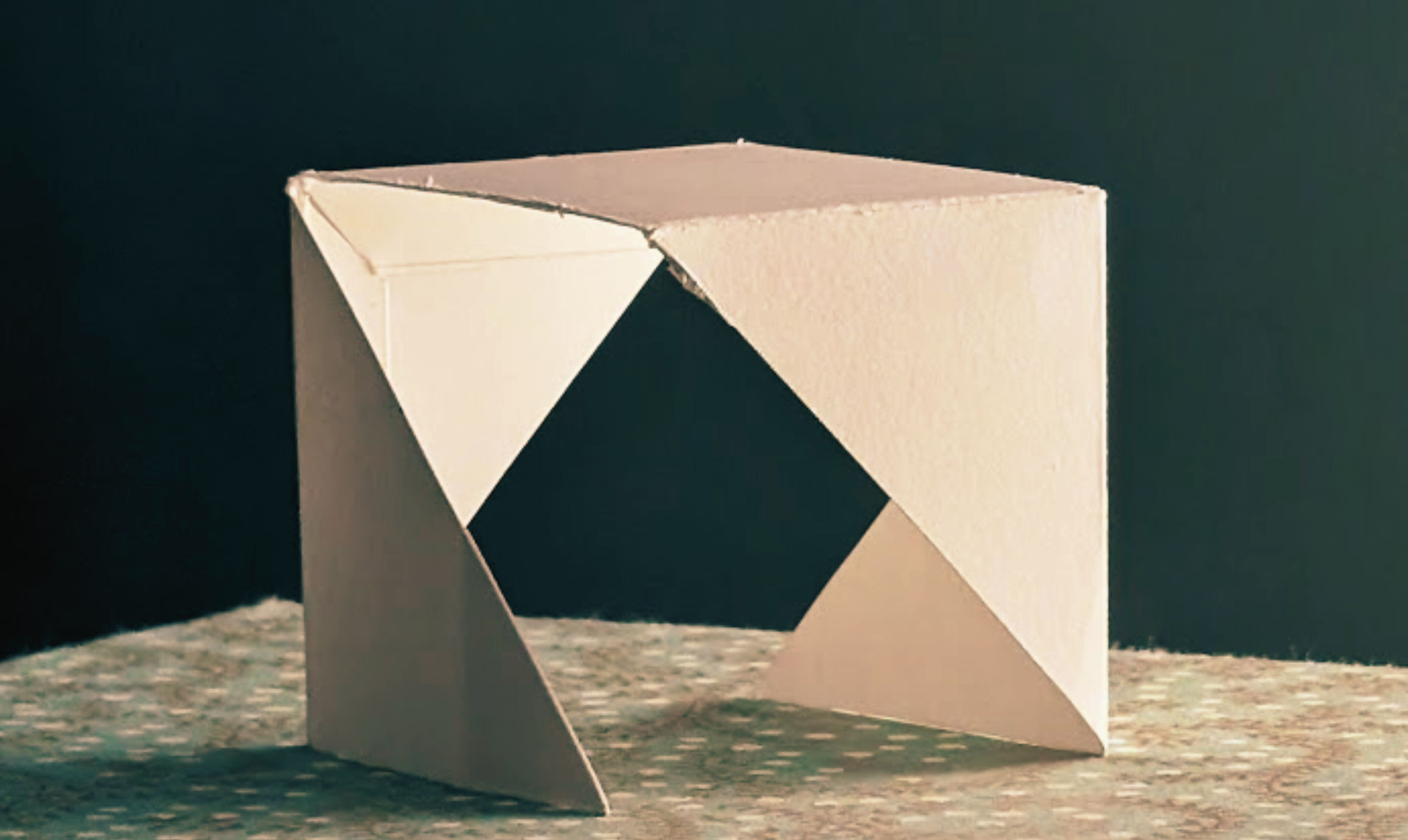 A modern geometric sculpture made of light-colored material, standing on a textured surface with a dark background.