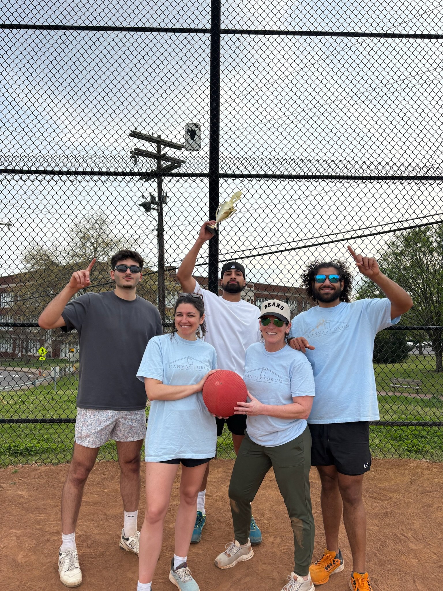 Group of six Canvas Homes teammates on a baseball field, celebrating after a game, some holding a trophy, with a fence and trees in the background.