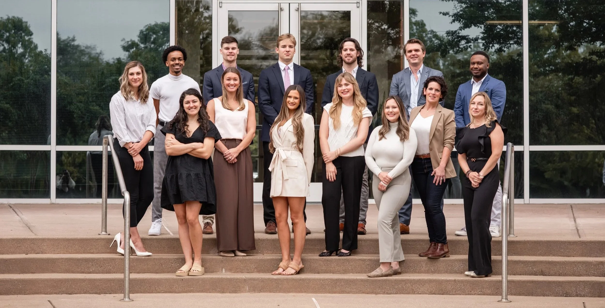 A diverse group of 15 people from the Canvas Homes team, both men and women, standing in front of a glass building with greenery reflected in the windows. They are dressed in business casual and formal attire, smiling on outdoor steps.