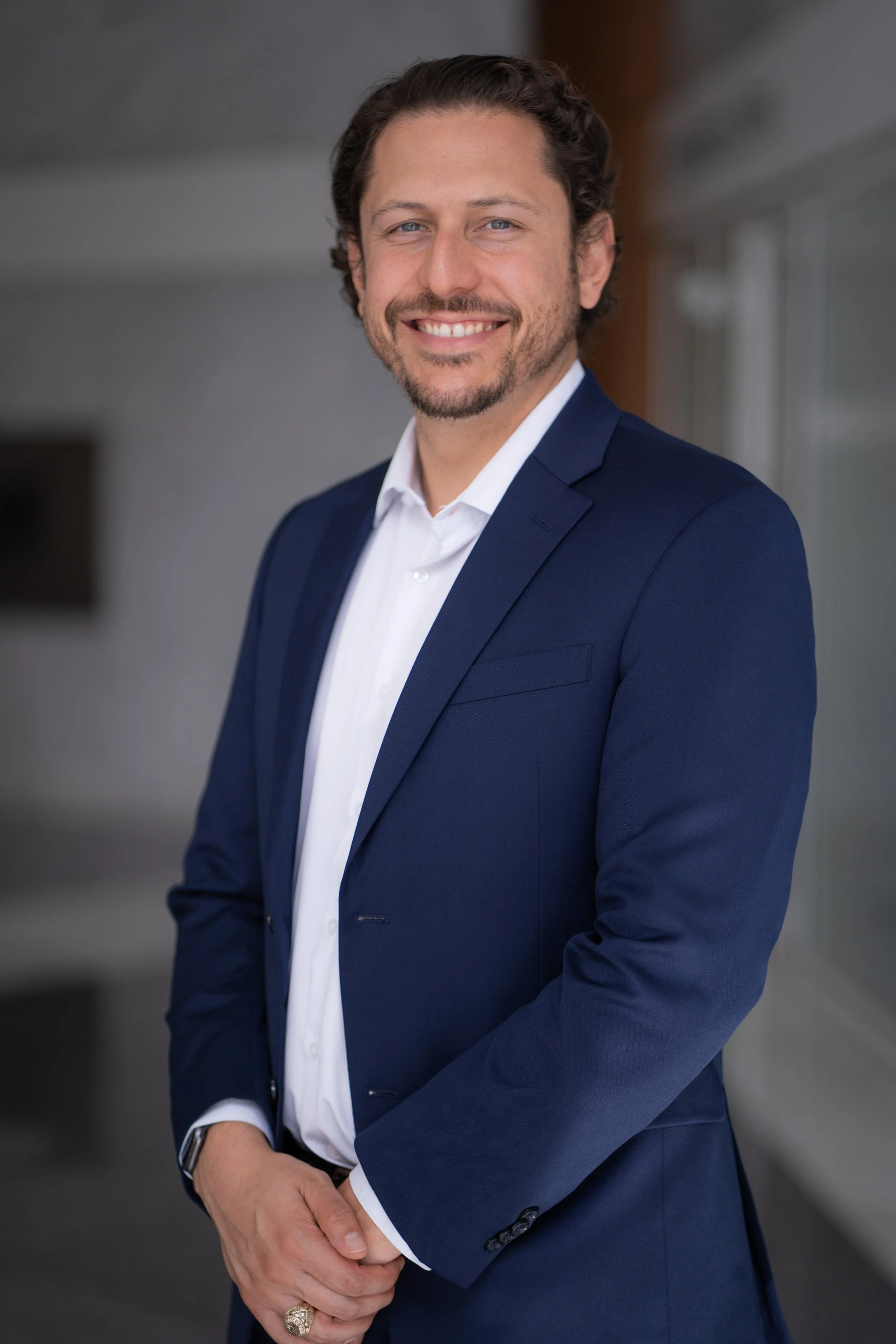 Portrait of a smiling man in a navy blue suit and white shirt, with his arms crossed, standing indoors with a neutral background.