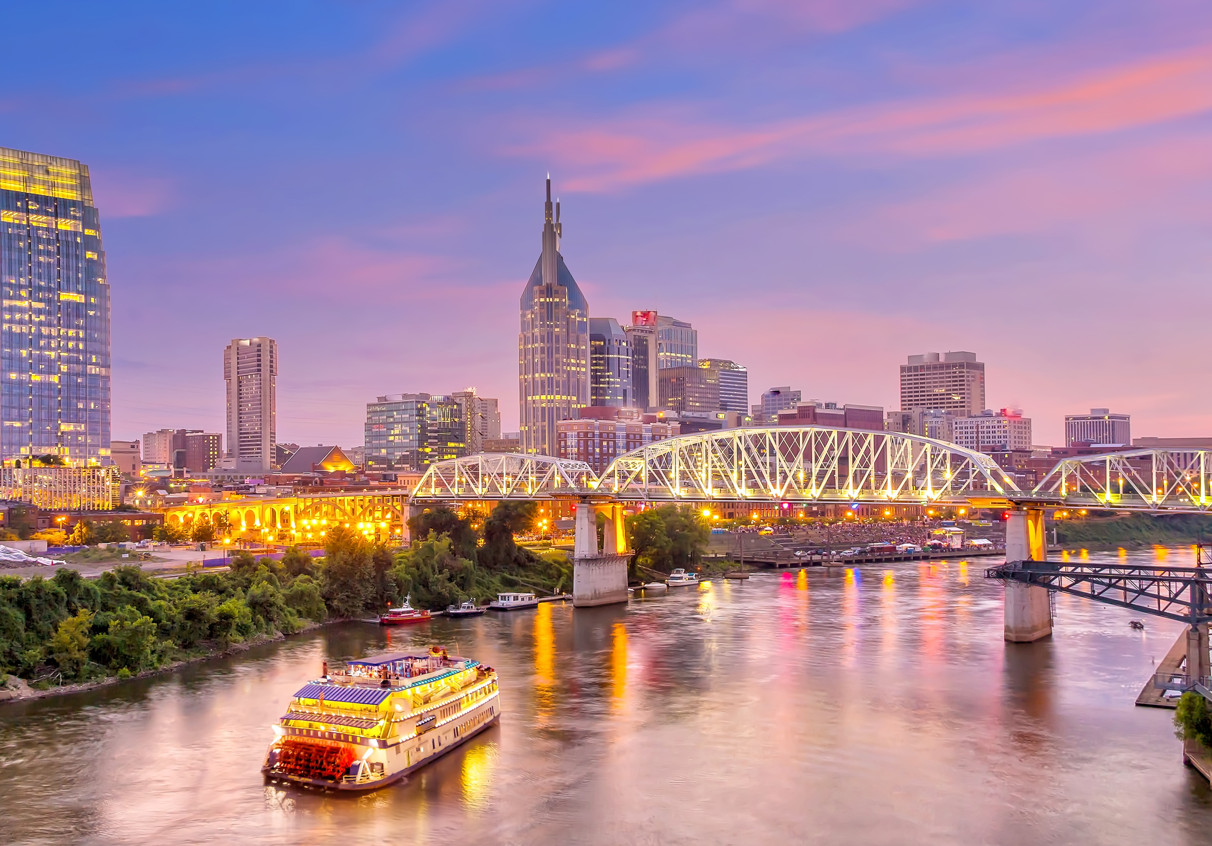 The Nashville, TN skyline at dusk with a river in the foreground, a cruise boat on the river, and skyscrapers with lit windows and a colorful sky with pink and purple clouds.