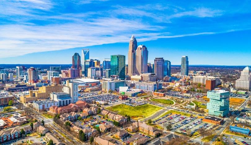 Aerial view of the Charlotte, NC skyline with tall modern skyscrapers, surrounded by residential areas and parking lots, under a partly cloudy blue sky.