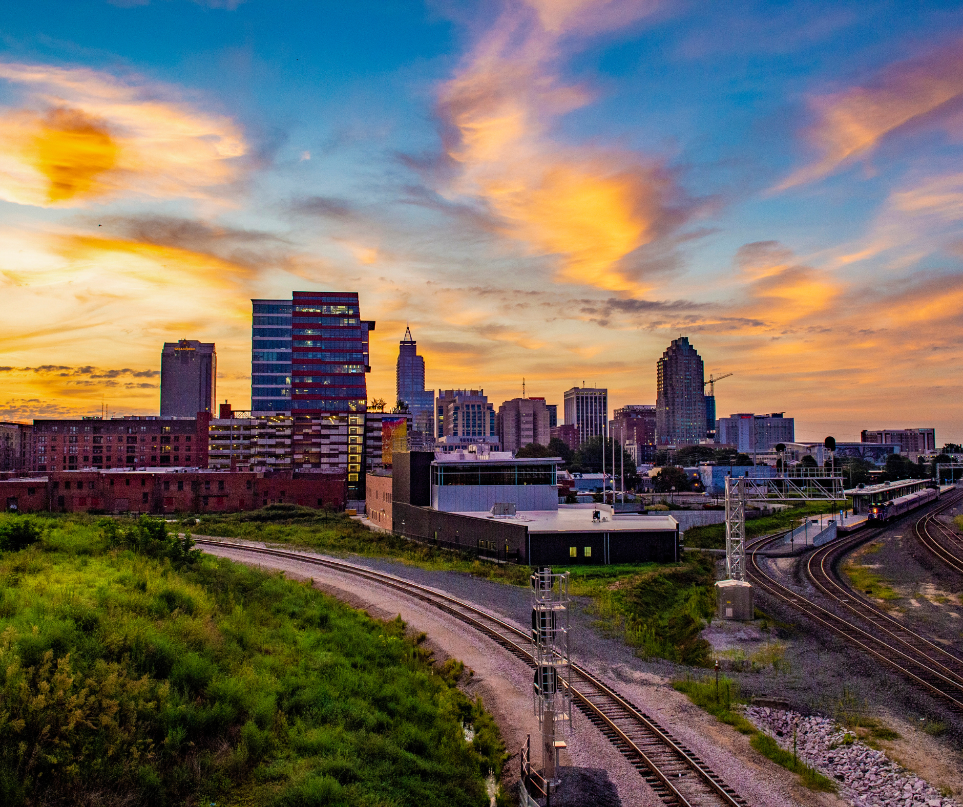 Sunset over the Raleigh, NC skyline with tall buildings and colorful clouds in the sky. Rails and greenery in the foreground.