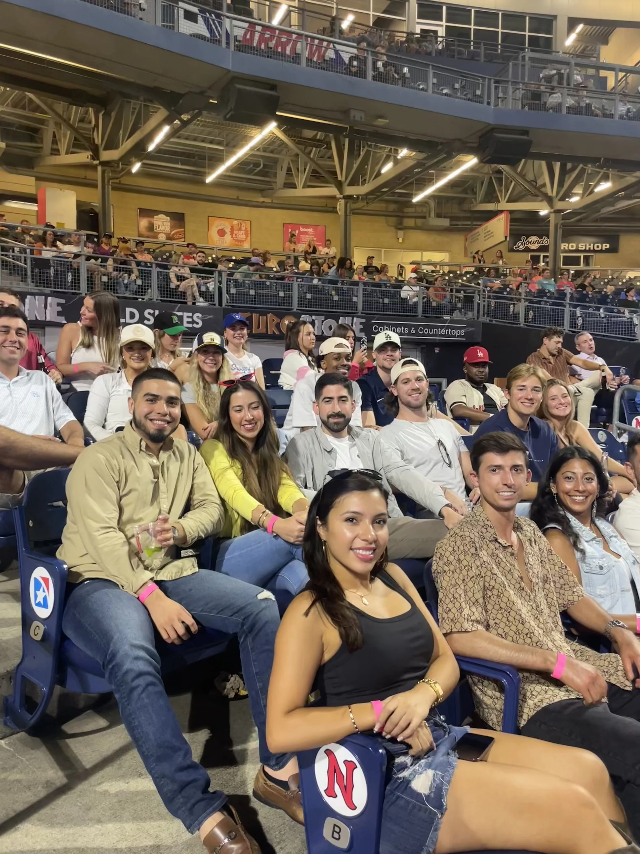 Crowd of people seated in stadium seats at a sports event, smiling and posing for the camera, with some wearing hats and casual clothing, in an indoor arena with multiple levels and bright lighting.