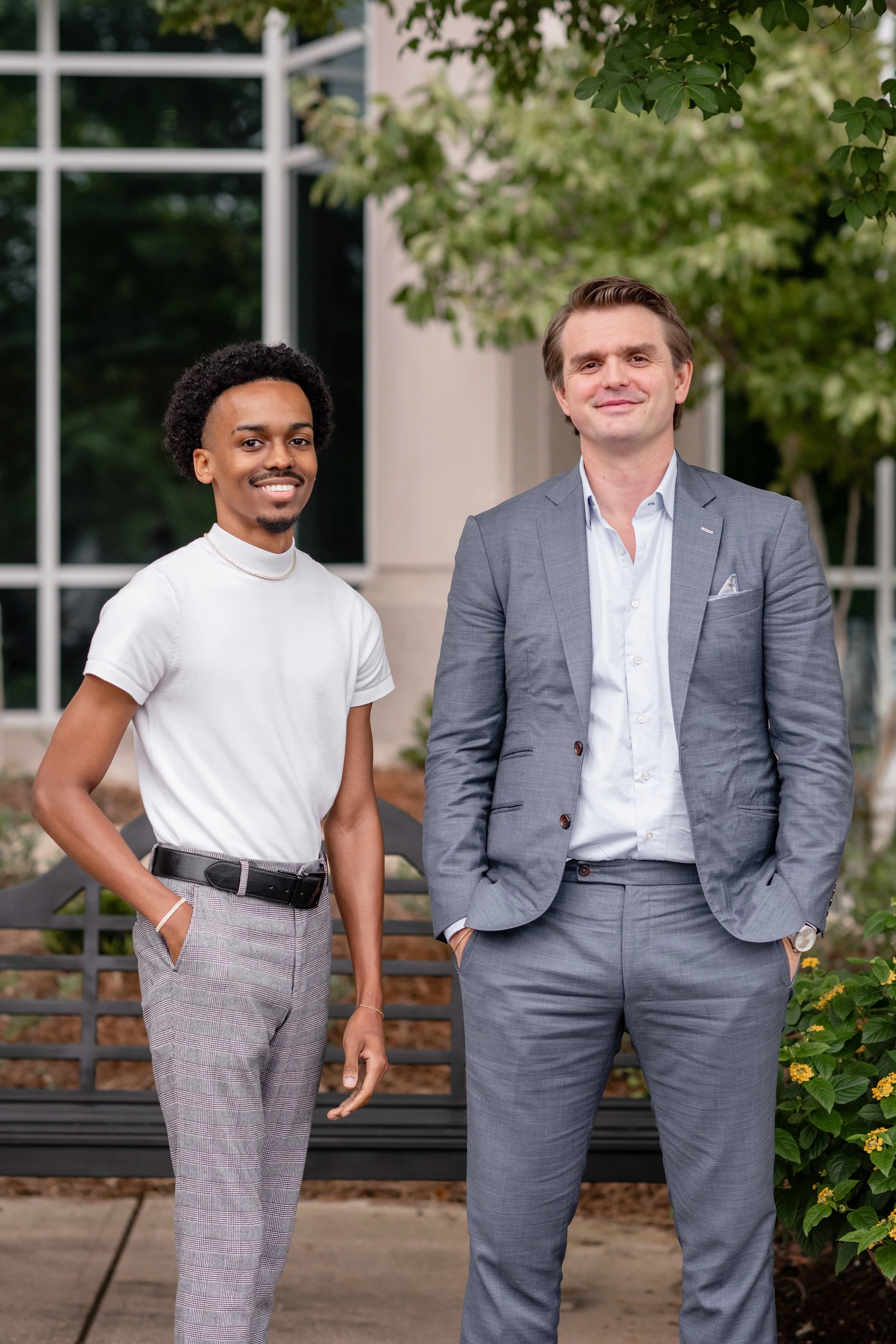Two professionally dressed men standing outdoors near a black park bench with greenery and trees in the background.