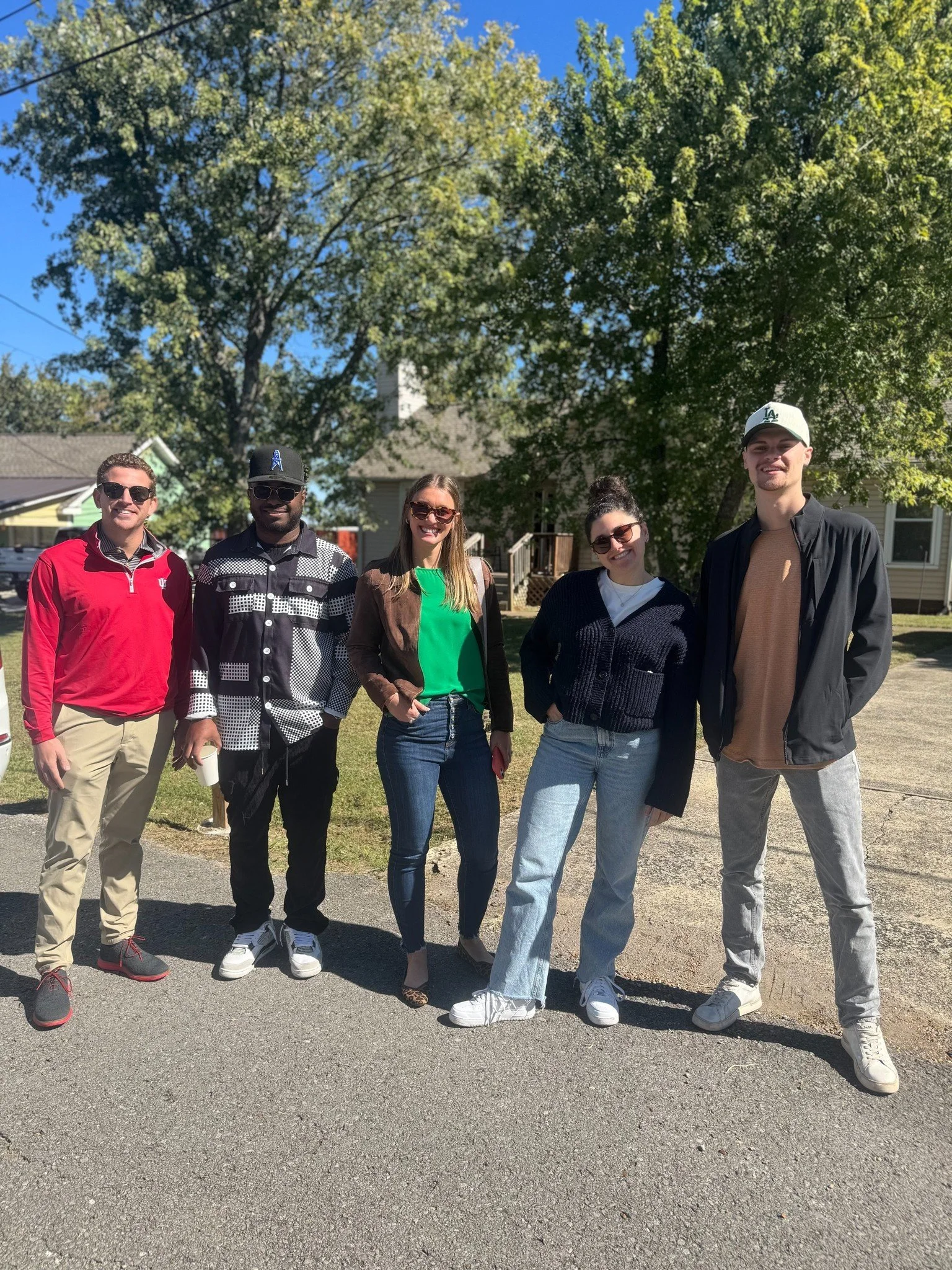 A group of five Canvas Homes teammates standing outdoors on a sunny day, smiling at the camera with a large tree and suburban houses in the background.