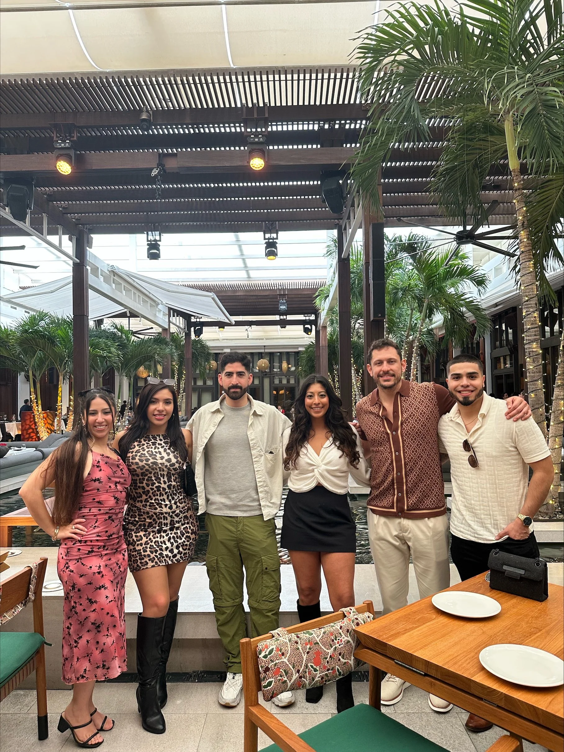Group of six Canvas Homes teammagtes smiling and posing for a photo in a restaurant with tropical plants and wooden decor. There are tables and chairs, some with decorative cushions, in the background.