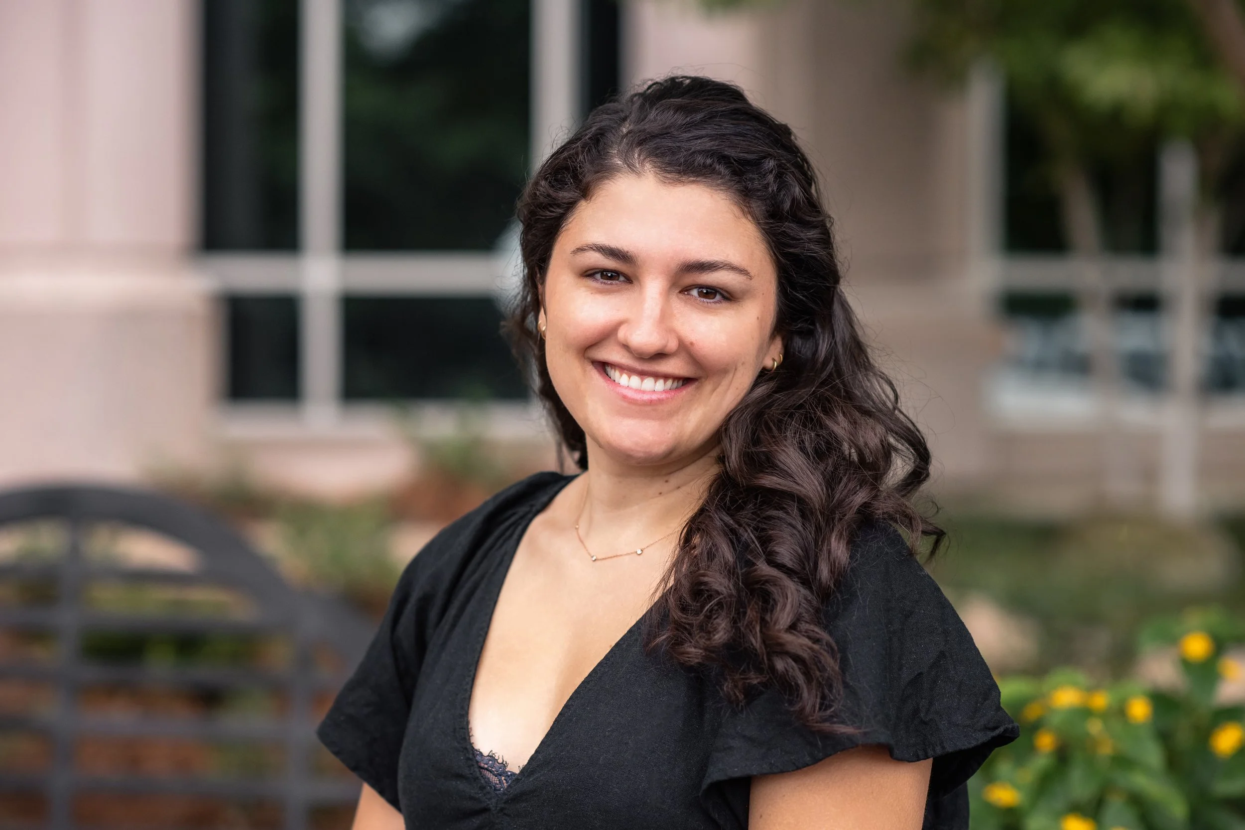 A smiling woman with long, dark, curly hair, wearing a black top and gold jewelry, standing outdoors near a park bench with background greenery and a building with large windows.