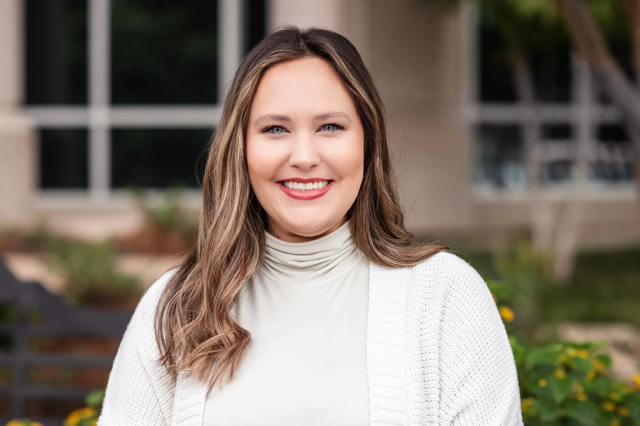 Portrait of a smiling woman with long wavy hair wearing a white turtleneck and cardigan, standing outdoors with blurred background of greenery and a building.