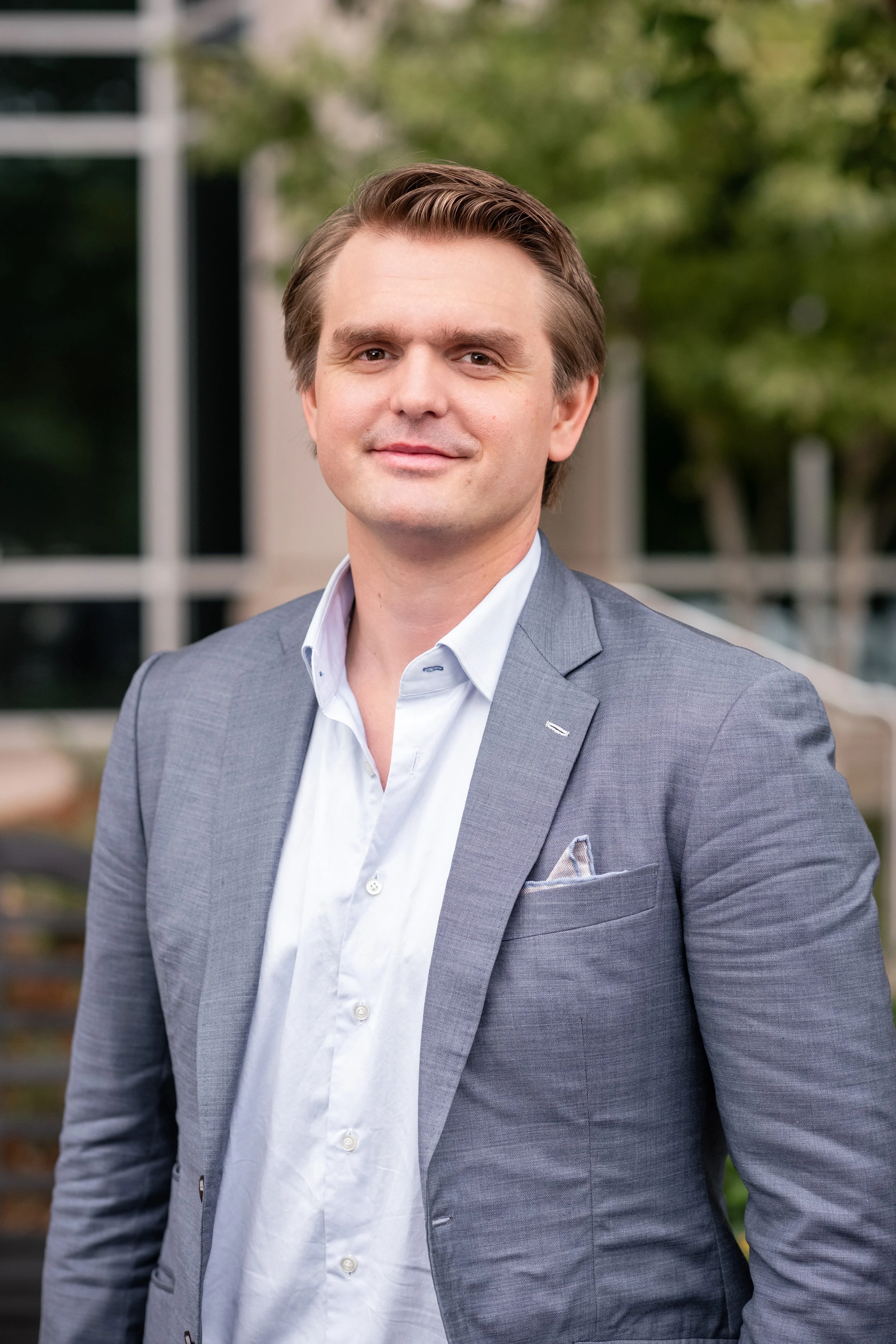Wyatt Tiffany, co-founder of Camvas Homes is in a grey suit and white shirt standing outdoors with green trees and a building in the background.