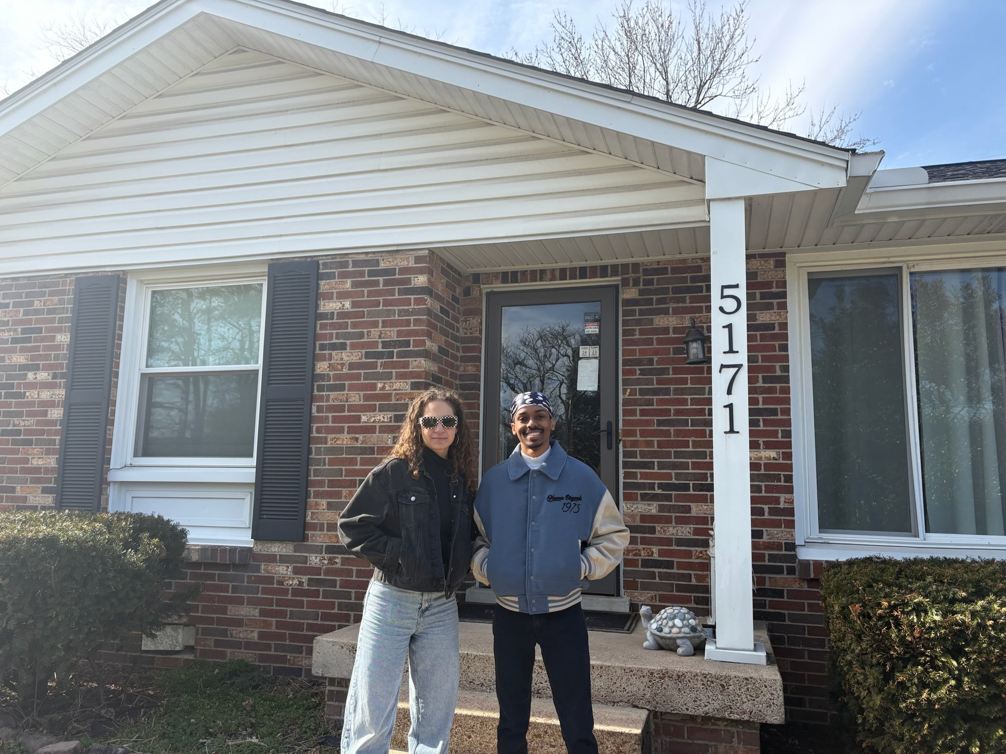 Two people are standing in front of a brick house with white siding, a front door, and a porch. The house has black shutters and a white pillar with the house number 5171. The individuals are smiling; the woman has long curly hair and is wearing sung