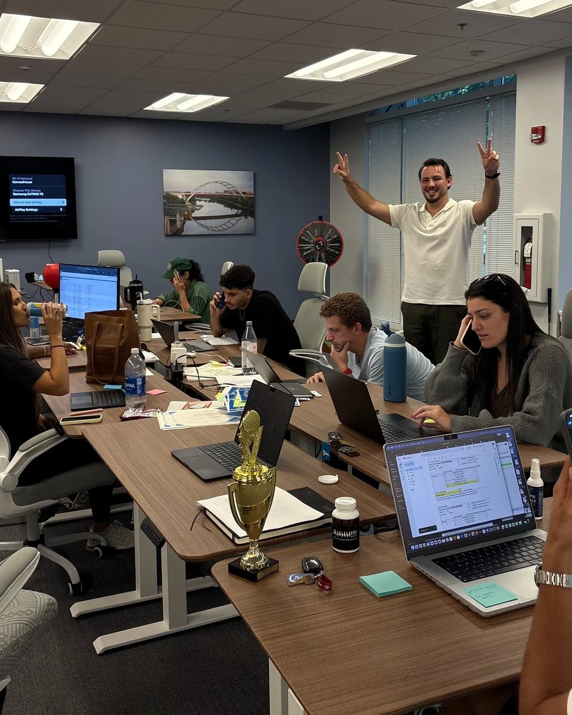 A group of people sitting around a conference table in a meeting room. One man stands at the back, smiling and making peace signs with both hands. Others are working on laptops or talking on the phone. There is a trophy on the table and a woman in th