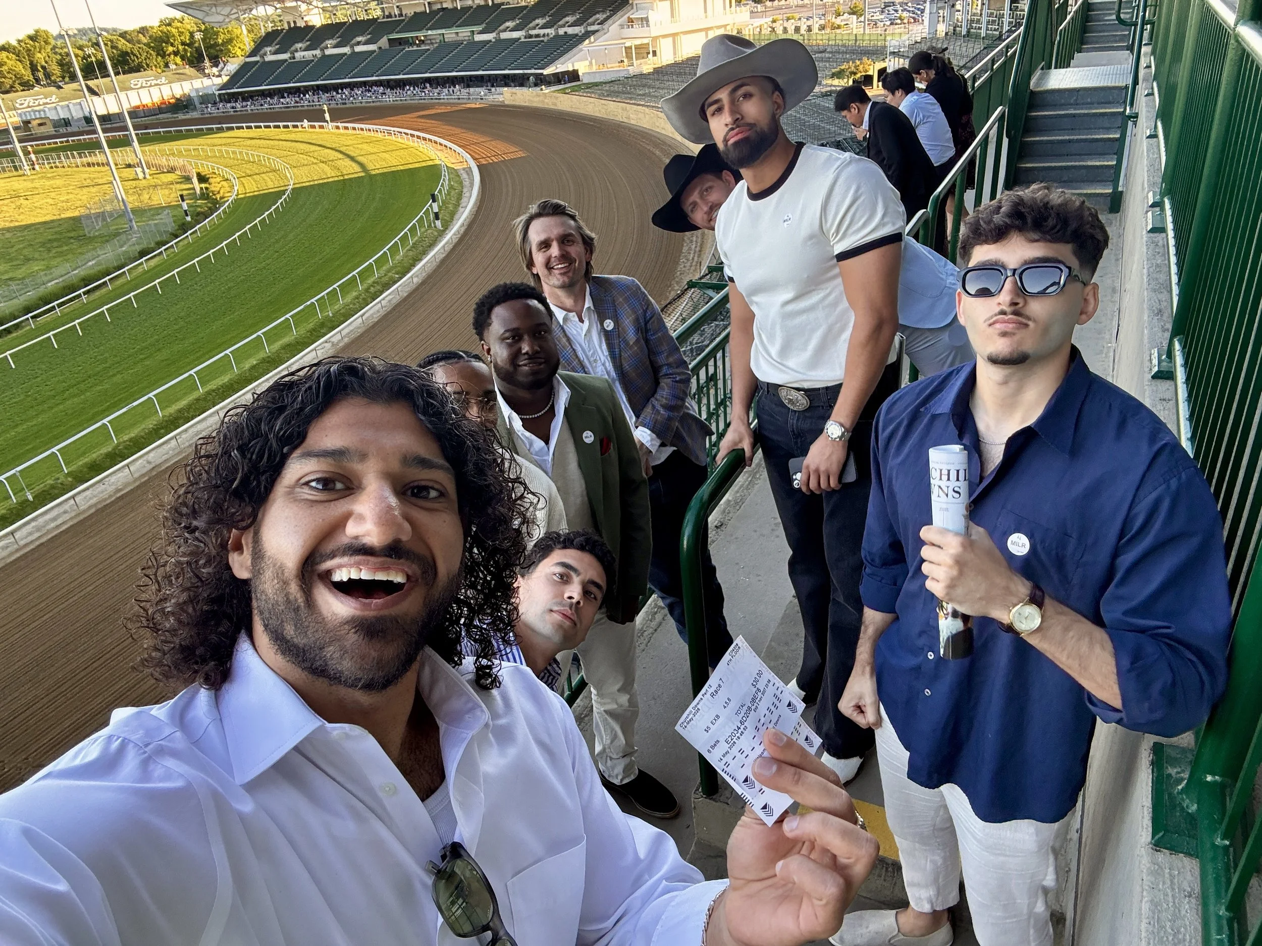 A group of Canvas Homes teammates at a horse racing track, some holding tickets, taking a selfie with the race track in the background, in the late afternoon.