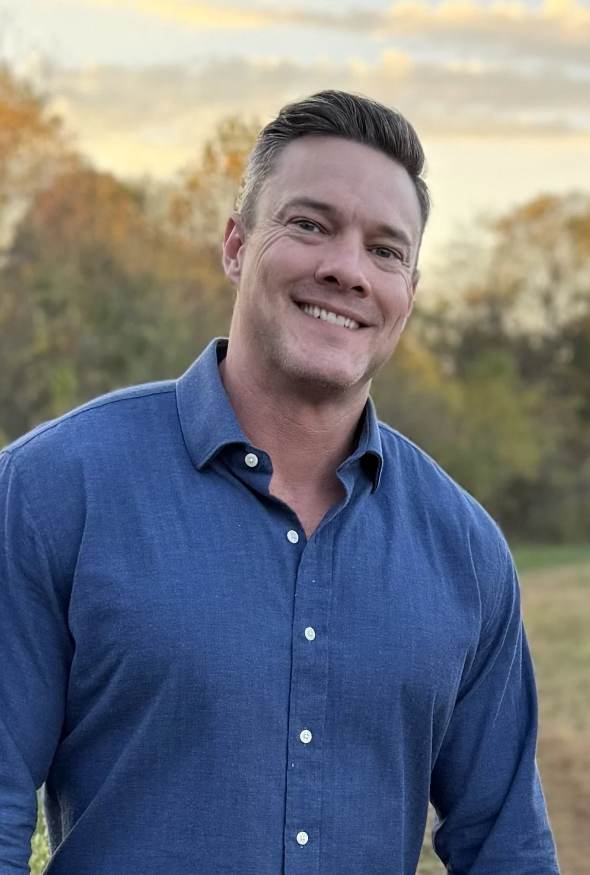 Mat Farmer, founder of Canvas Homes, is smiling and wearing a blue button-up shirt. He is outdoors with autumn trees and a cloudy sky in the background.