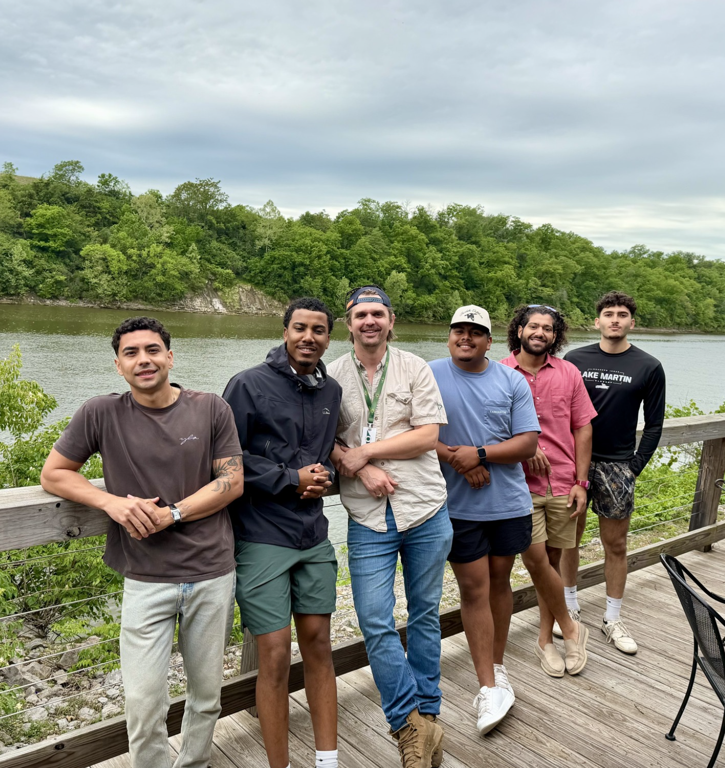 A group men from the Canvas Home team are standing on a wooden deck near a body of water with lush green trees in the background, overcast sky.