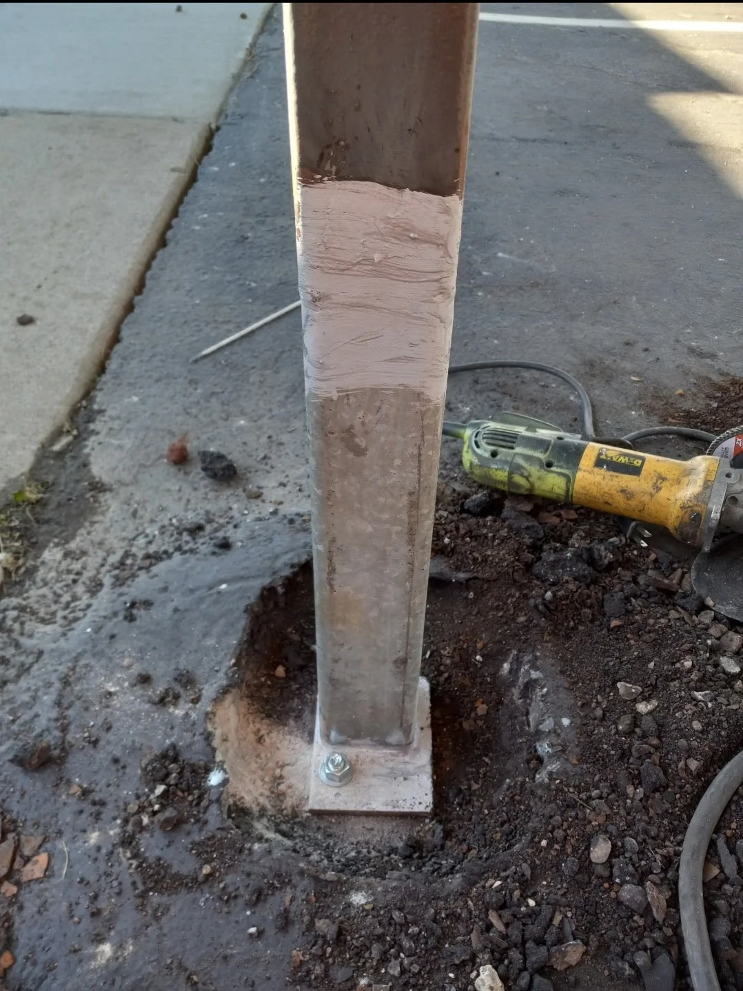 A metal post secured to the ground with a bolt, with a power drill lying nearby on the dirt, on a sidewalk.