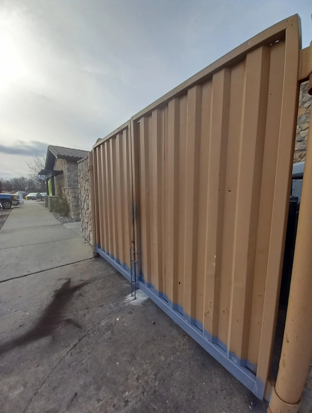 A tall wooden gate with vertical planks and metal framework, situated beside a stone building, with a sidewalk and parked cars visible in the background.