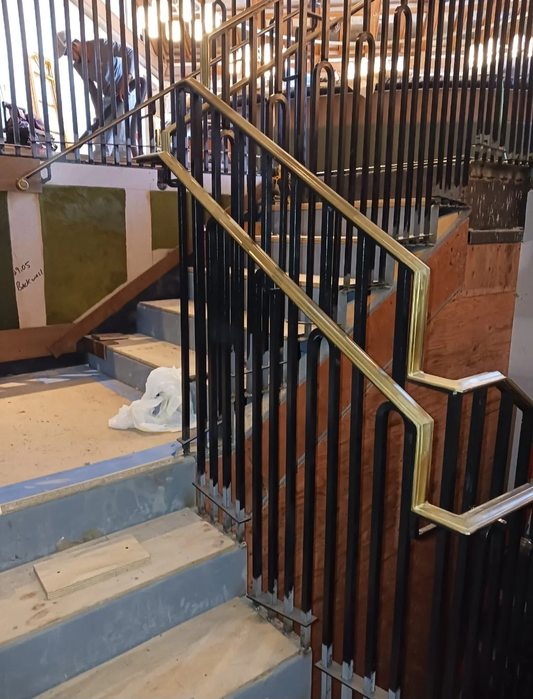 A staircase under construction with black metal railings and a wooden section, with a person working in the background.