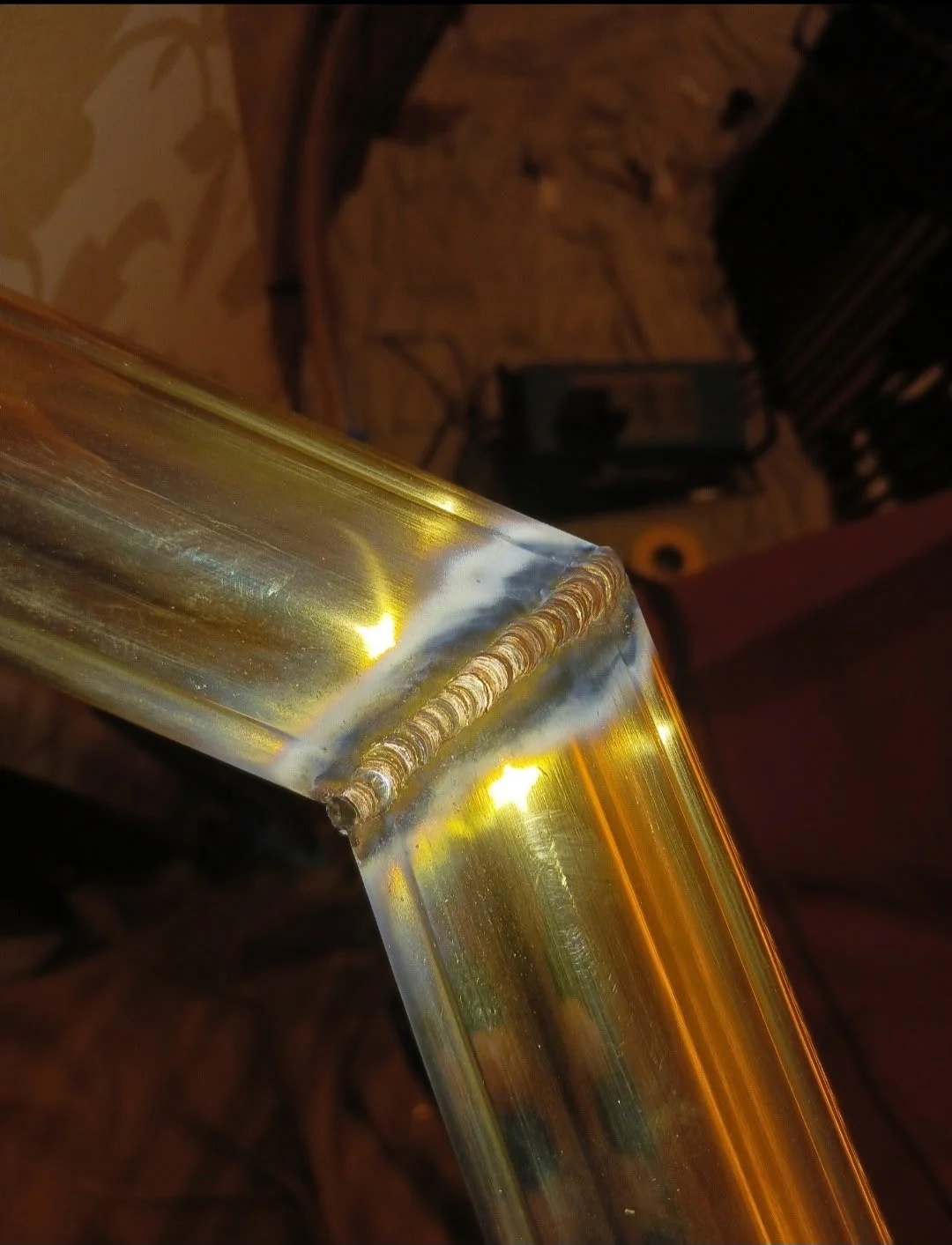 Close-up of a welded metal joint on a shiny, reflective golden-colored metal pipe or bar, with a workshop background including tools and equipment.
