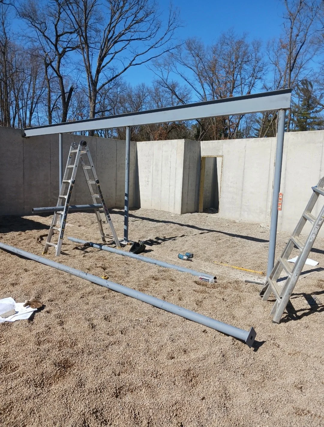 Construction site with metal frame, ladders, and concrete walls, set on gravel ground under clear blue sky.