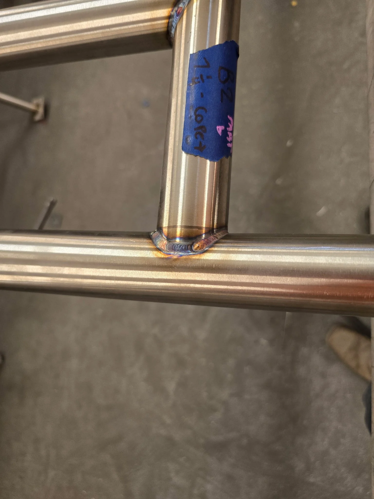 Close-up of a welded metal joint on a tubular metal frame with a blue identification label.