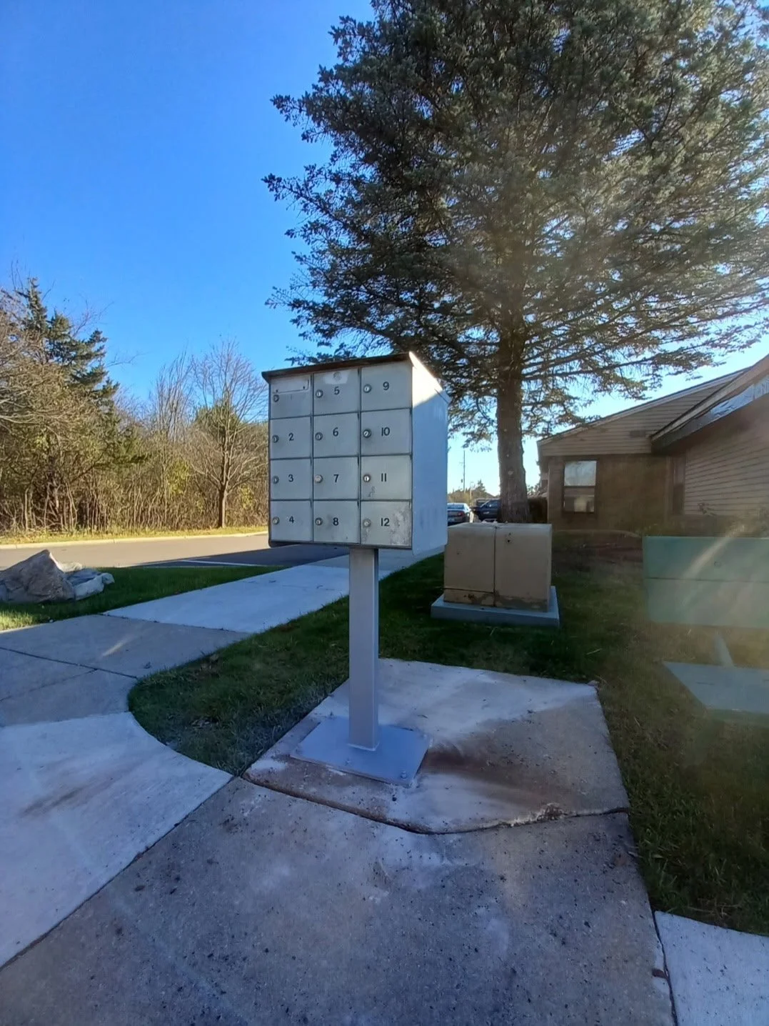An outdoor mailbox station with twelve individual lockers arranged in a 3x4 grid, located on a sidewalk near trees and a residential building.