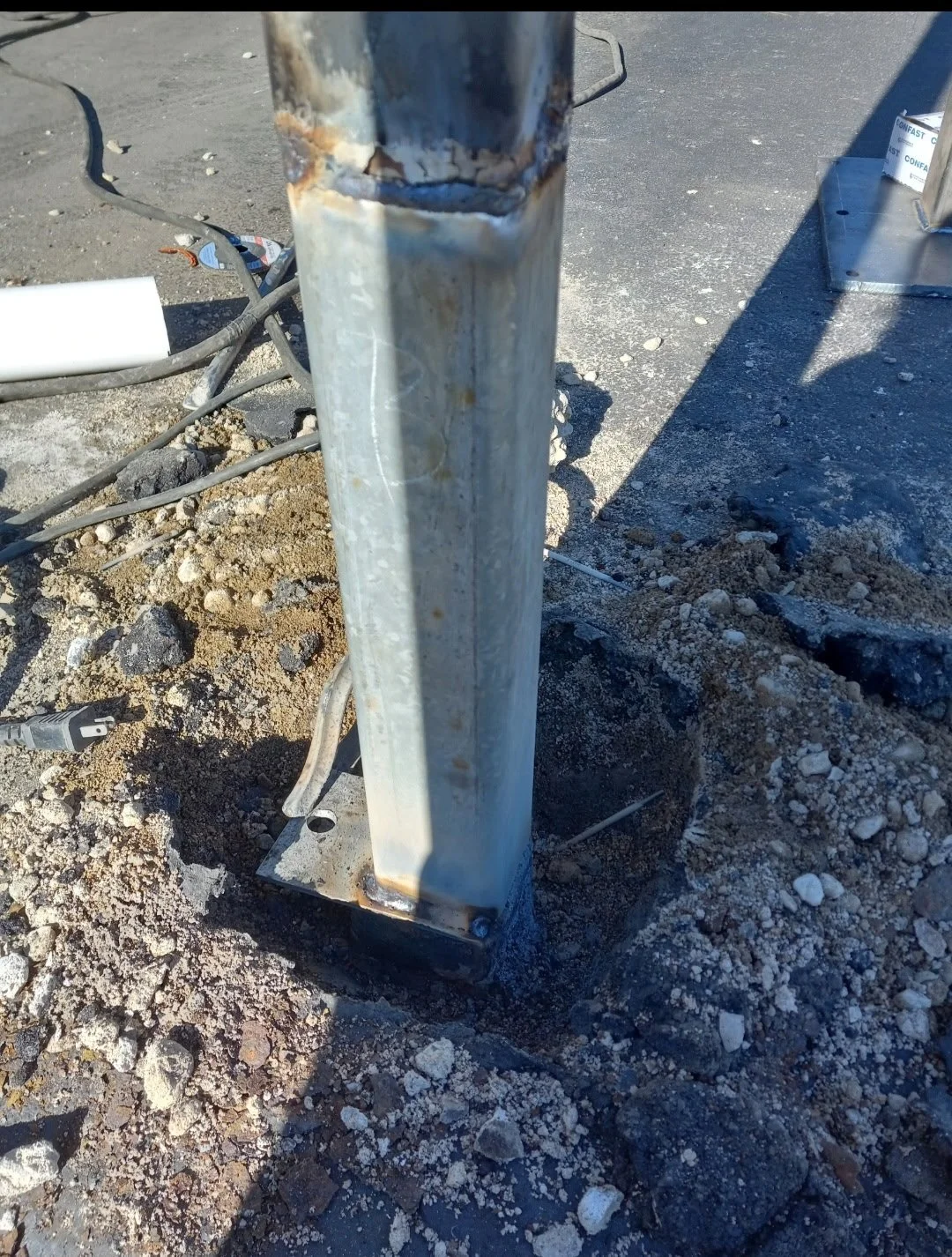 Close-up of a metal support pole with signs of rust, installed on a construction site with dirt, rocks, and exposed wires around it.