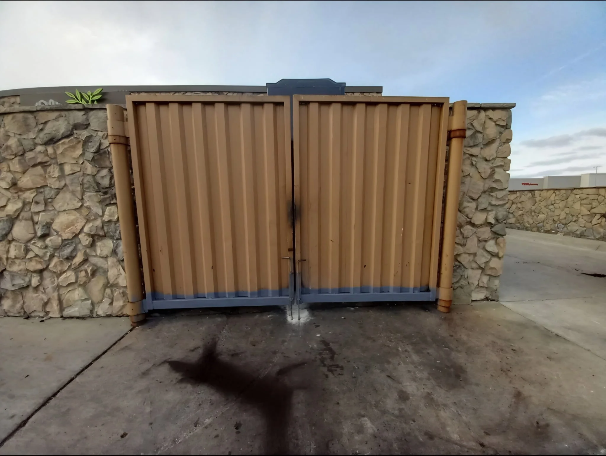 A tan metal gate with vertical ridges, surrounded by stone walls on both sides, with a concrete ground in front and a blue sky in the background.