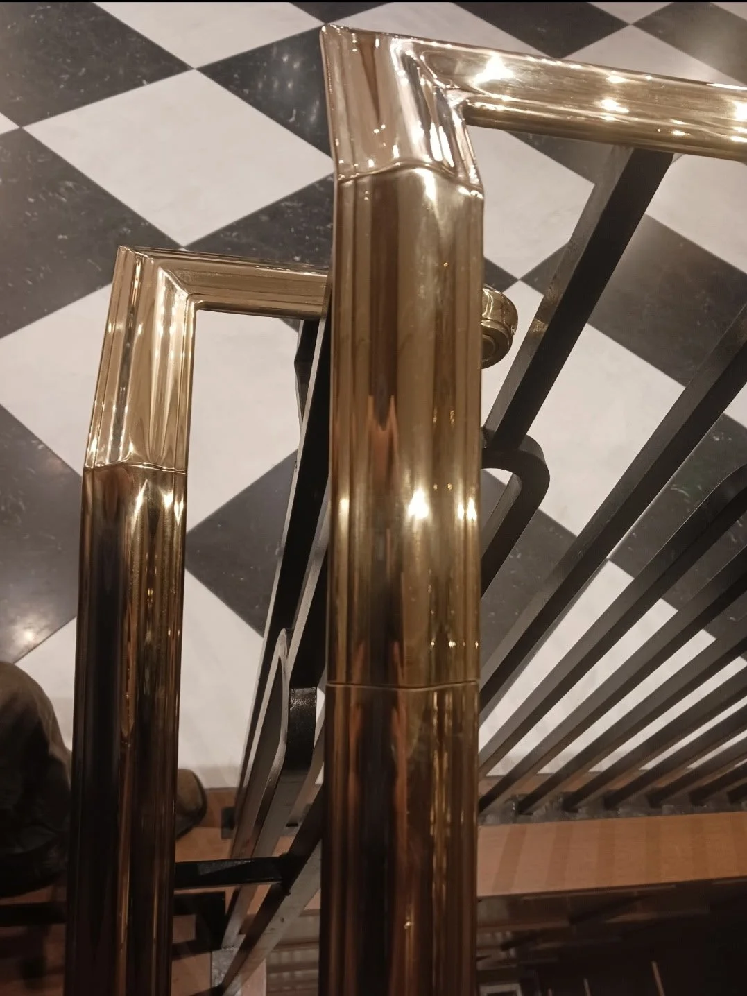 Top view of a polished gold-colored metal handrail on a staircase with black vertical bars, overlooking a black and white checkered tiled floor.