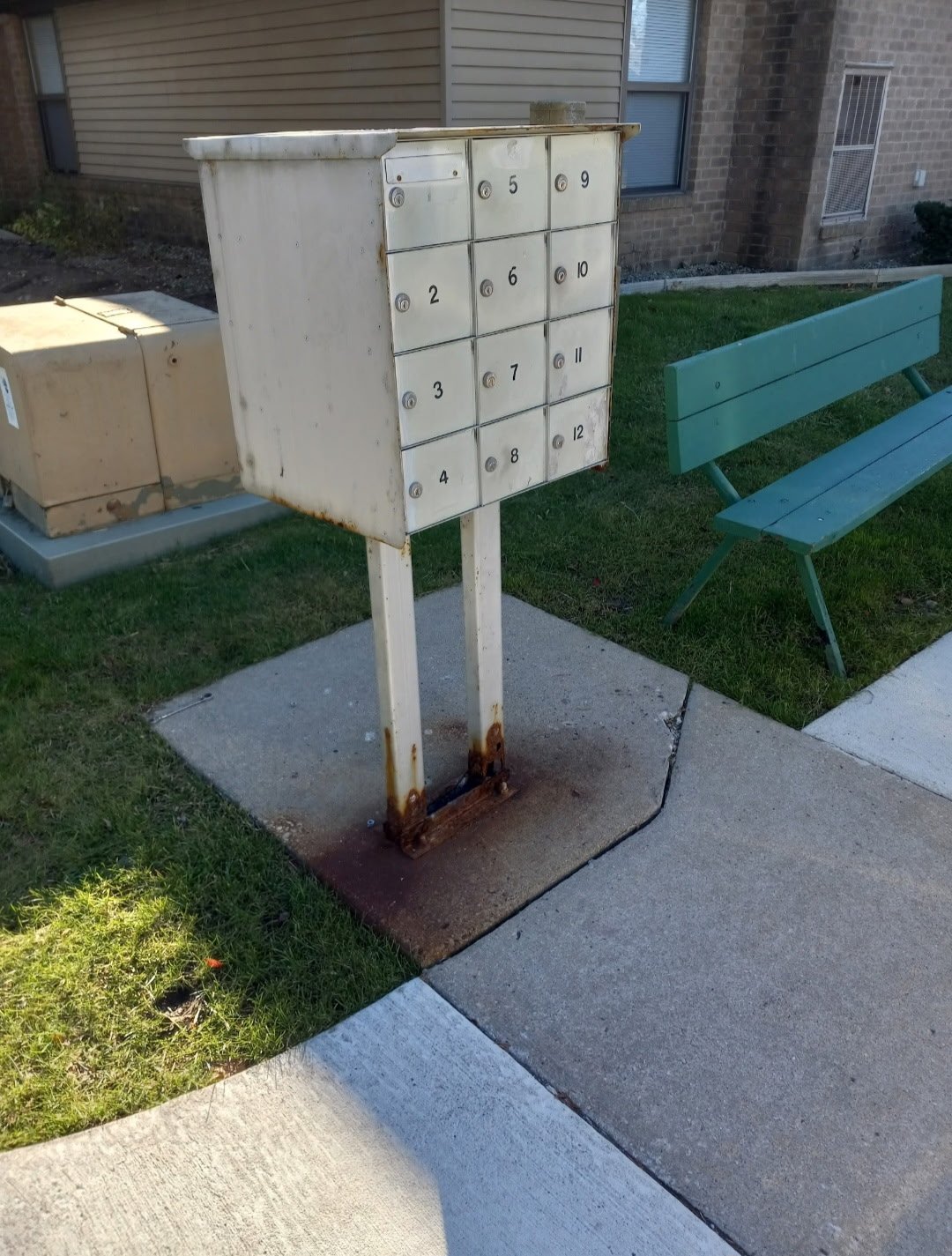 A white metal mailbox with 12 compartments numbered 1 to 12, mounted on two metal legs with rust at the base, located on a sidewalk near a green park bench and residential building.