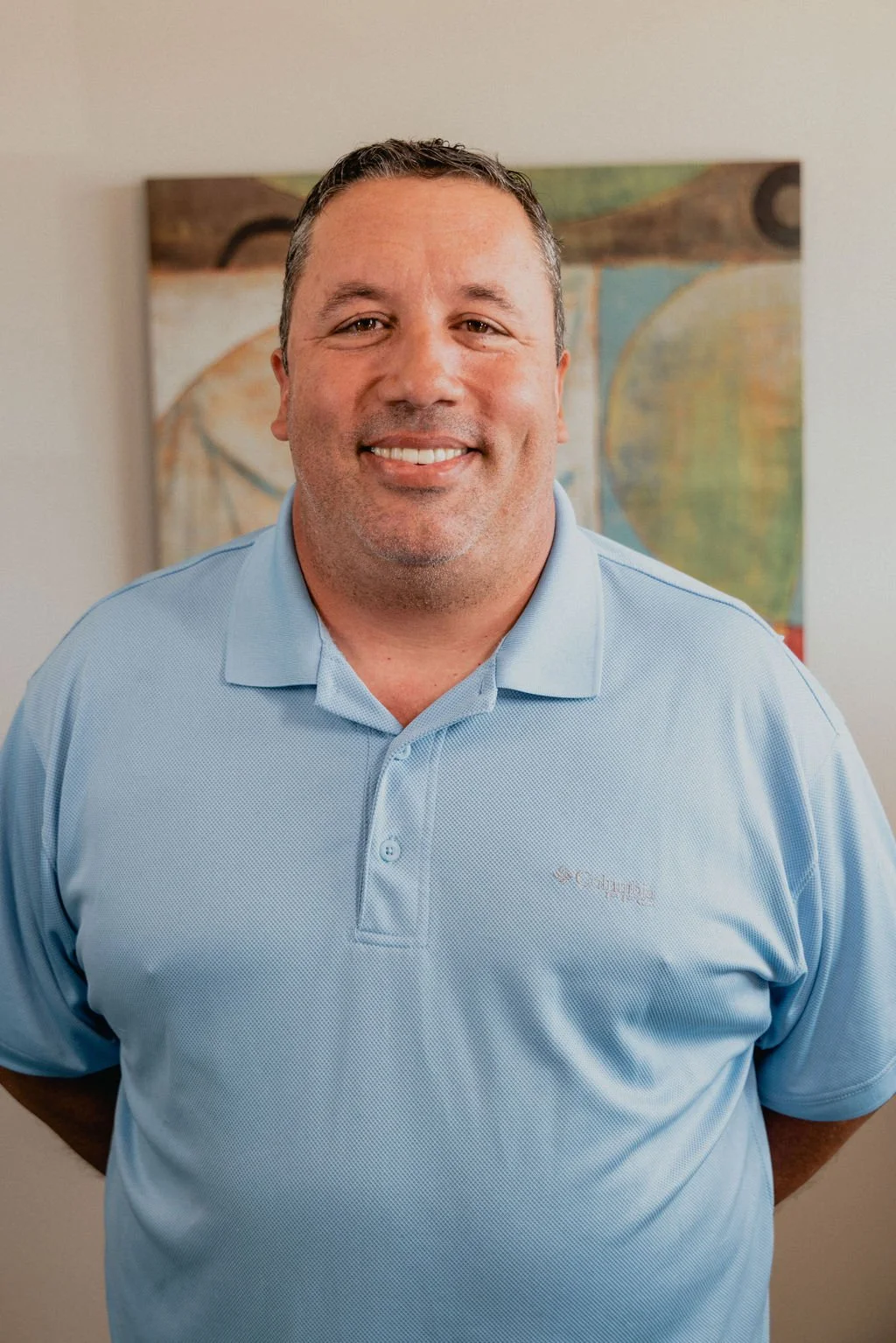 A smiling man with short dark hair, wearing a light blue polo shirt, standing in front of an abstract painting on a plain wall.