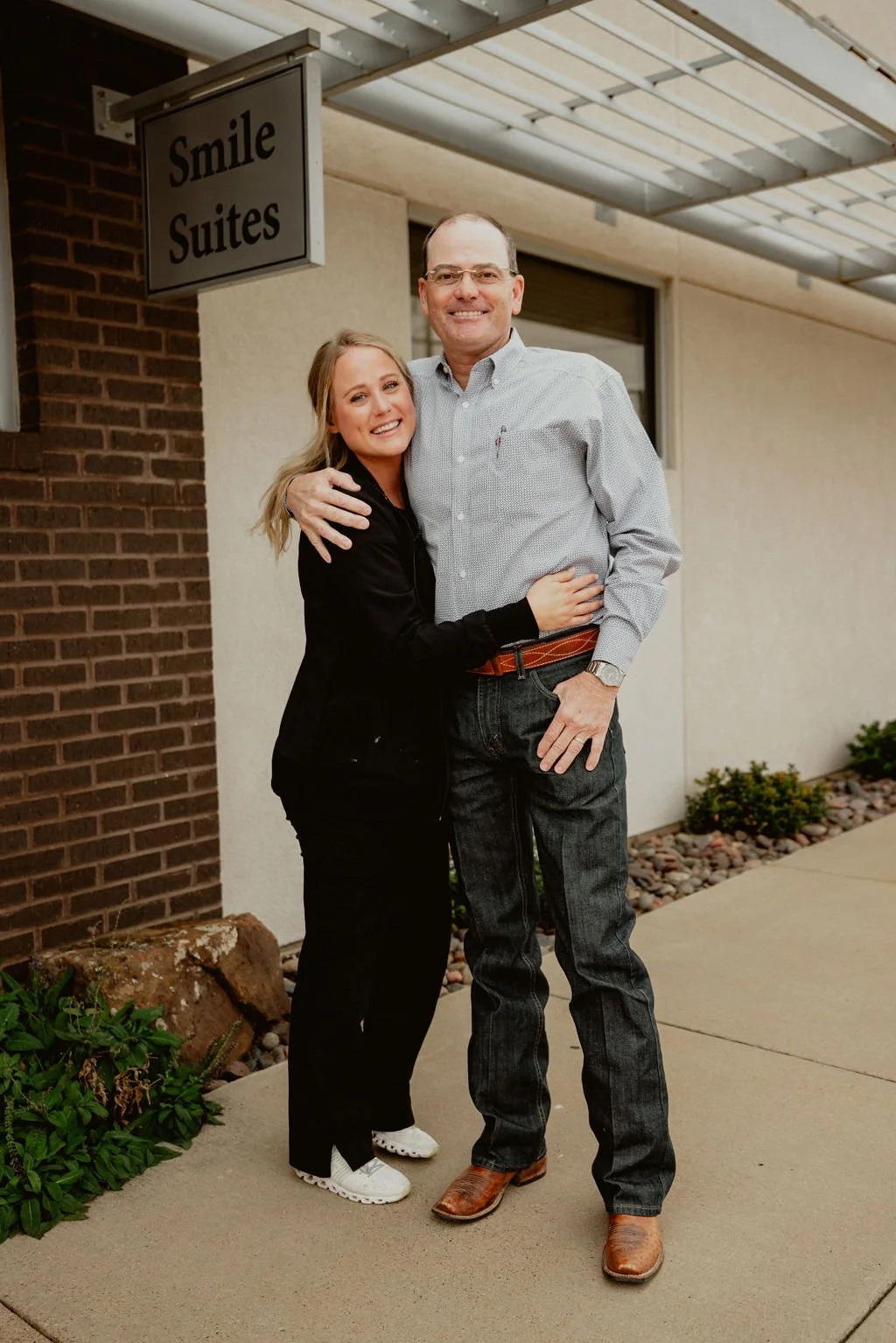 A young woman and an older man standing outside a building with a sign that reads 'Smile Suites'. They are smiling, hugging, and seem happy. The woman is wearing black pants, a black jacket, and white sneakers. The man is dressed in jeans, a light gr