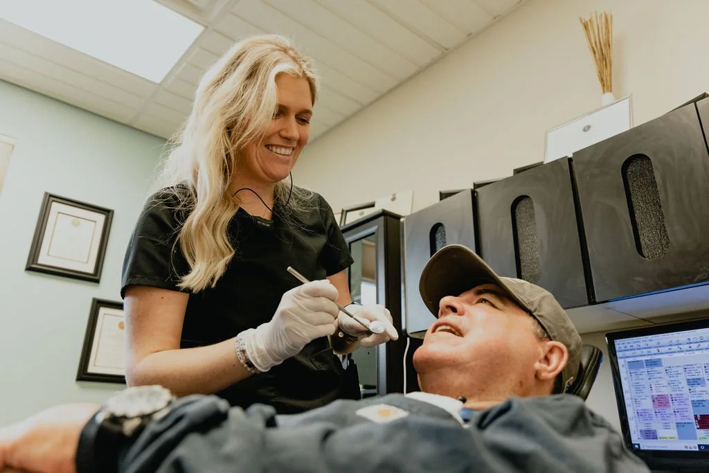 A woman with blonde hair and gloves smiling while giving a dental injection to a man in a cap, seated in a dental office.