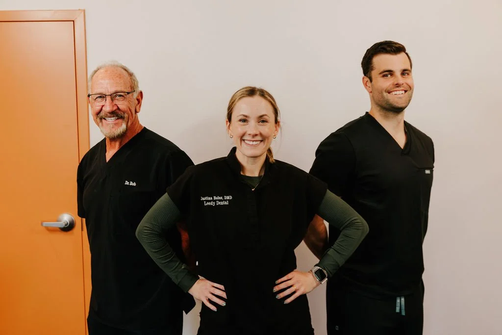 Three dental professionals standing against a plain wall, smiling at the camera.