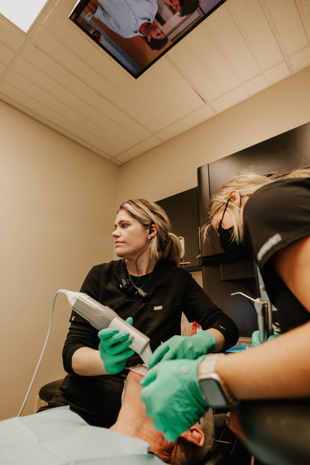 Dental professionals perform a procedure on an elderly patient's mouth in a medical room with a mounted television screen above.