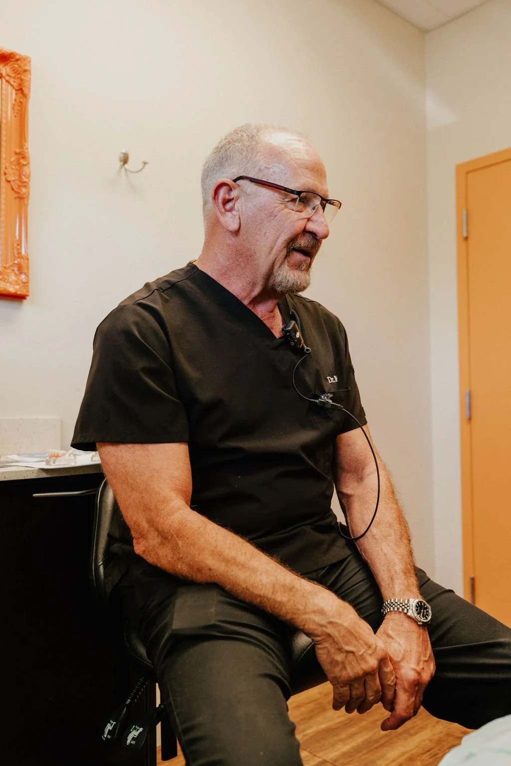 A middle-aged man with gray hair, beard, and glasses, wearing a black medical uniform, sitting on a chair in a room with cream-colored walls, orange door, and artwork on the wall.