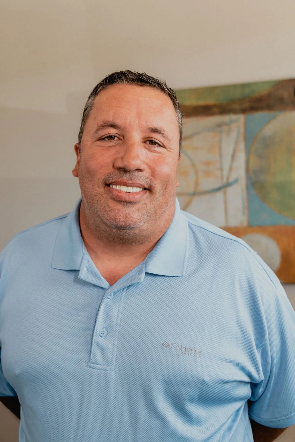 A smiling man with short dark hair and a stubbled beard wearing a light blue polo shirt standing in front of an abstract painting.