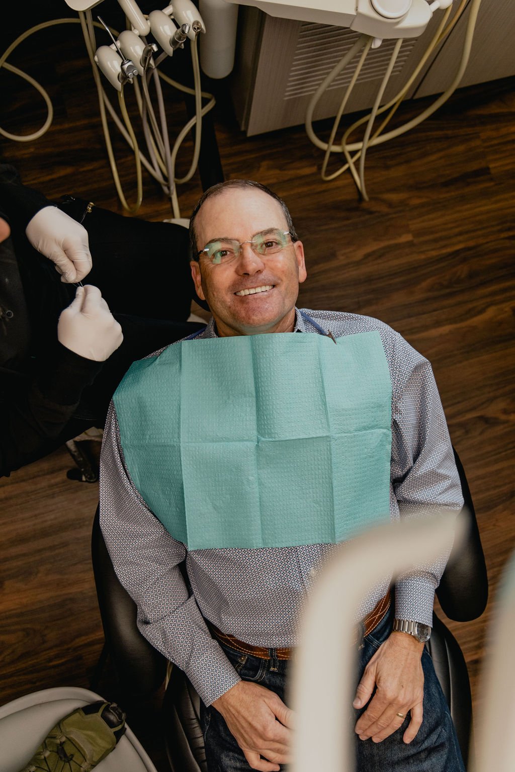 A man with glasses and a patterned shirt smiling while sitting in a dental chair, with a blue dental bib on his chest. A dental professional, wearing gloves, is preparing dental tools nearby in a dental office.