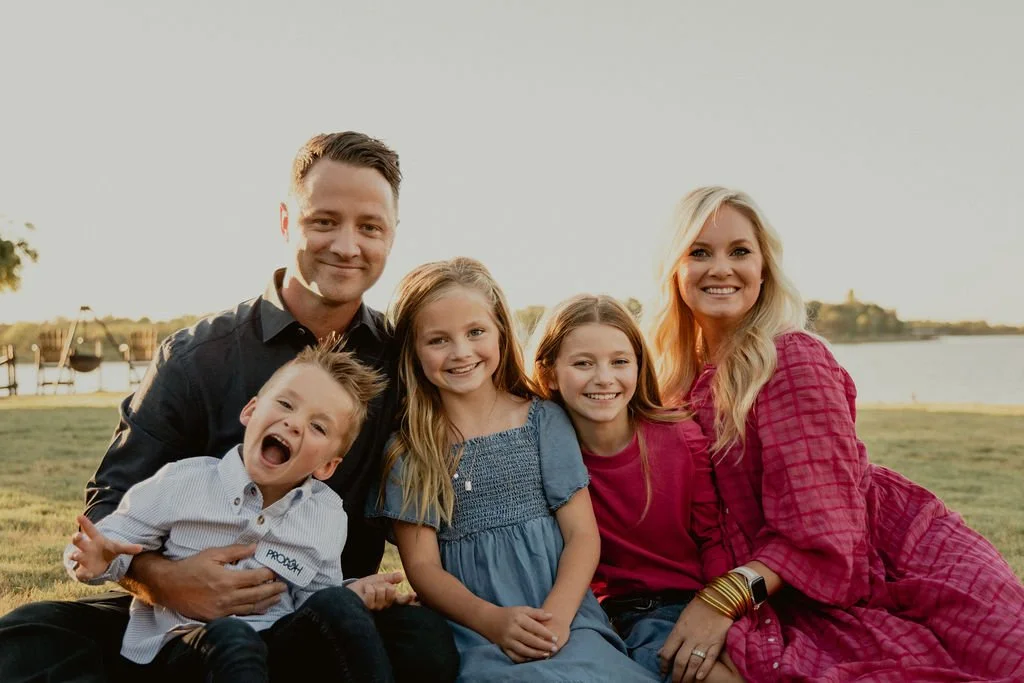 A family of five sitting on the grass near a body of water during sunset, smiling at the camera.