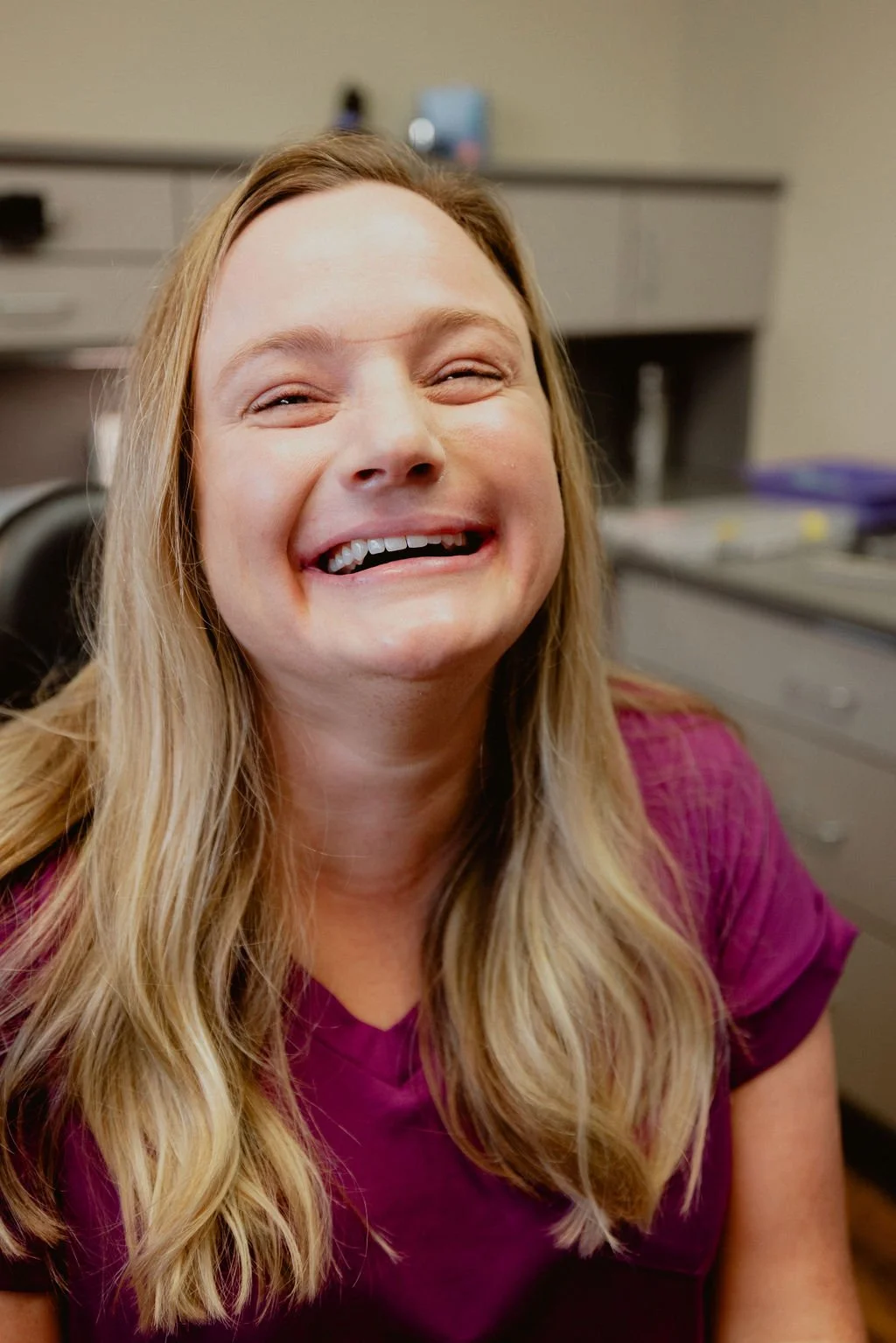Close-up of a woman with long blonde hair, smiling broadly, wearing a purple top, in an office setting with cabinets and supplies in the background.