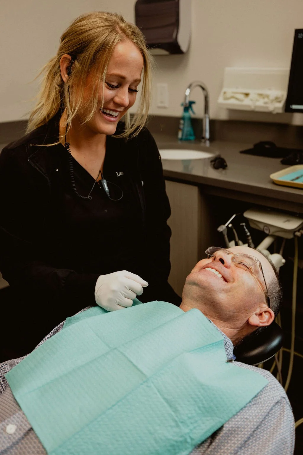 A dental professional smiling and looking at a patient lying in a dental chair with a green bib, in a dental clinic setting.