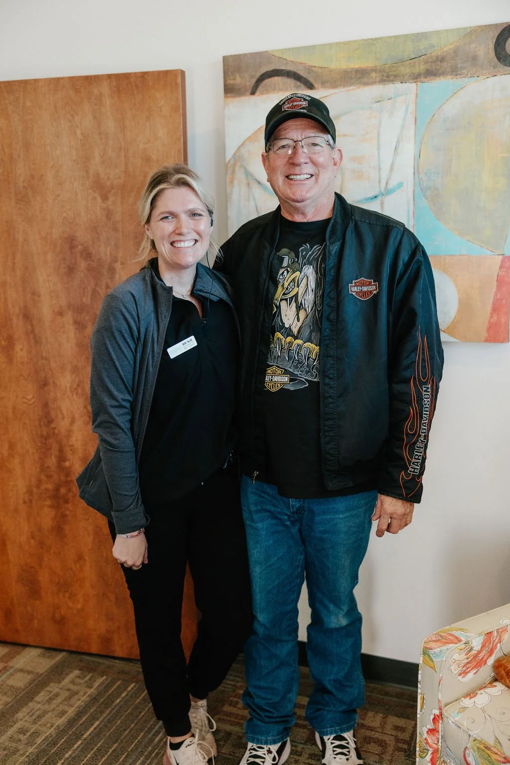 A woman and a man are standing side by side indoors, smiling for the camera. The woman has blonde hair, wears a black shirt, black pants, and a gray jacket with a name tag. The man has glasses, short hair, and is wearing a black Harley Davidson jacke