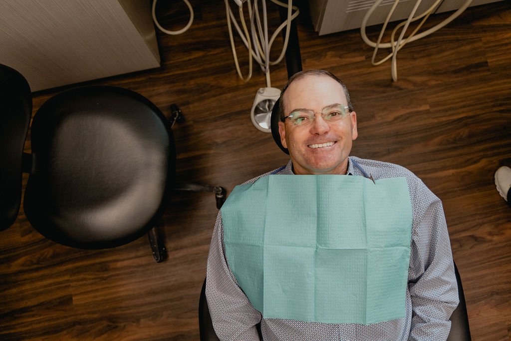 A man with glasses and a blue button-down shirt, smiling, sitting in a dental chair with a green dental bib, in a dental clinic with wooden flooring and dental equipment in the background.